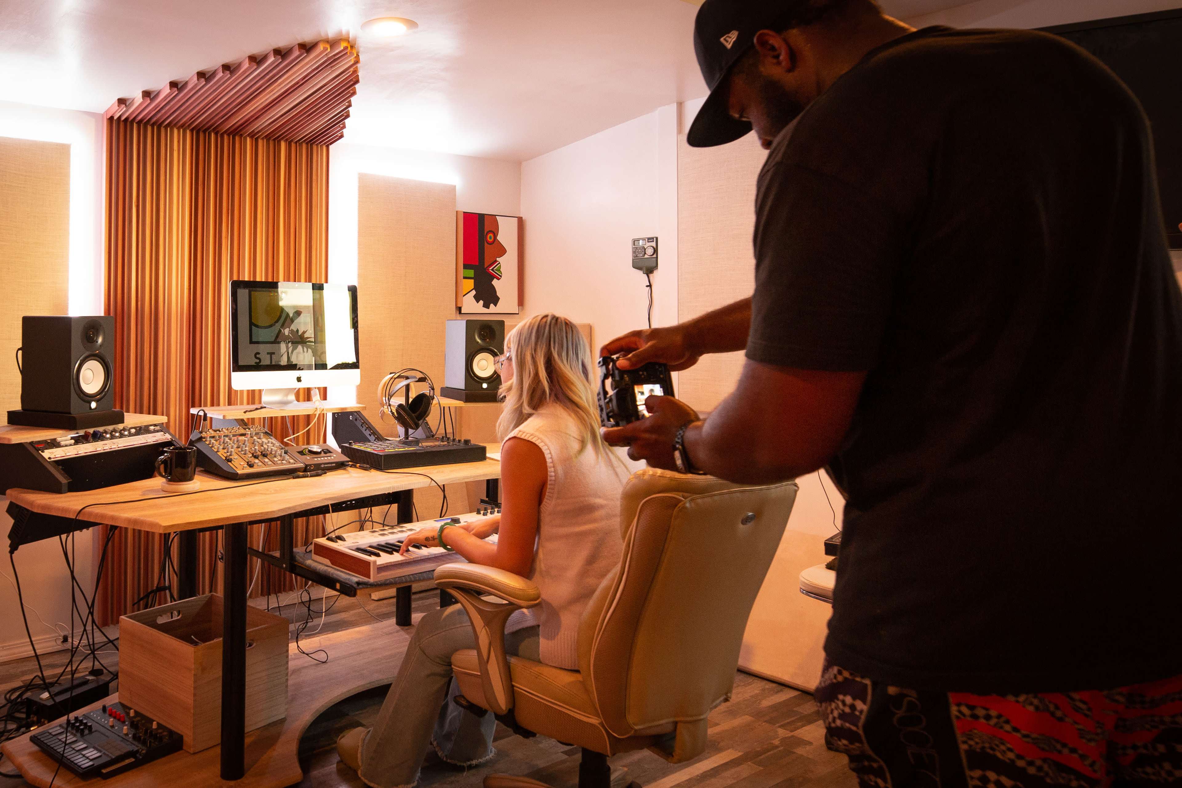 A person sits at a keyboard in a recording studio while another person takes a photo of them.