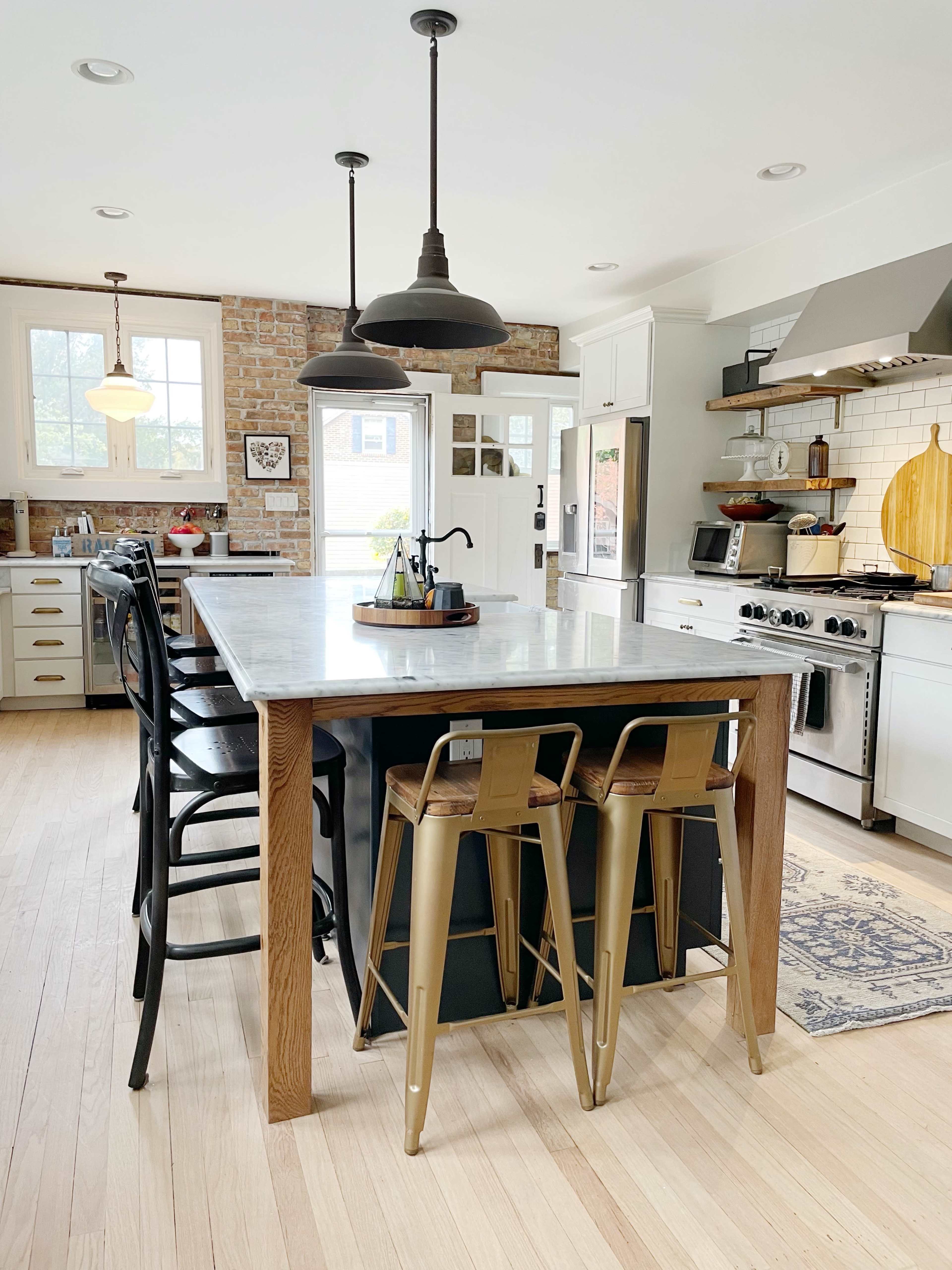 The kitchen features a large island with a marble countertop, black and gold bar stools, and stainless steel appliances, set against exposed brick and white cabinetry.