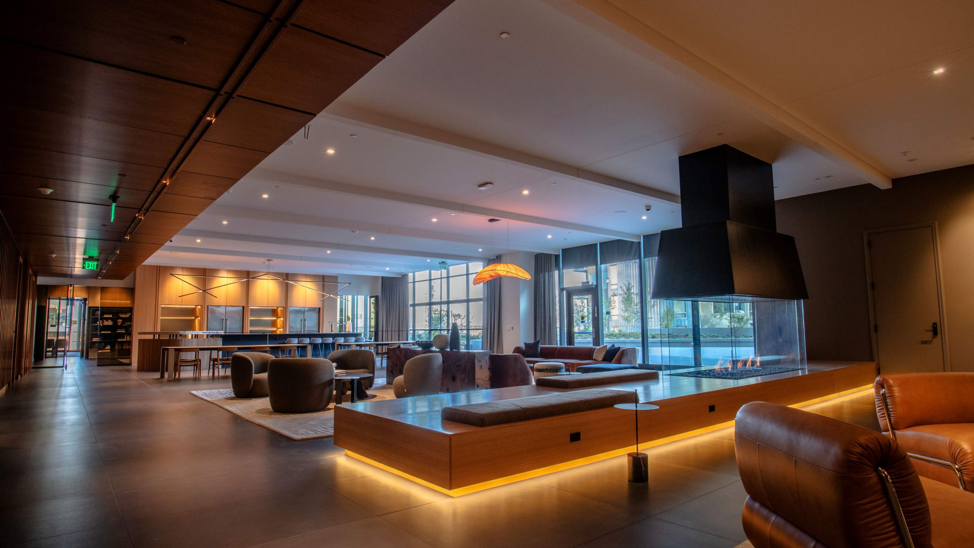 The image shows a modern lounge area with wooden accents, comfortable seating, and large windows allowing natural light to fill the space.