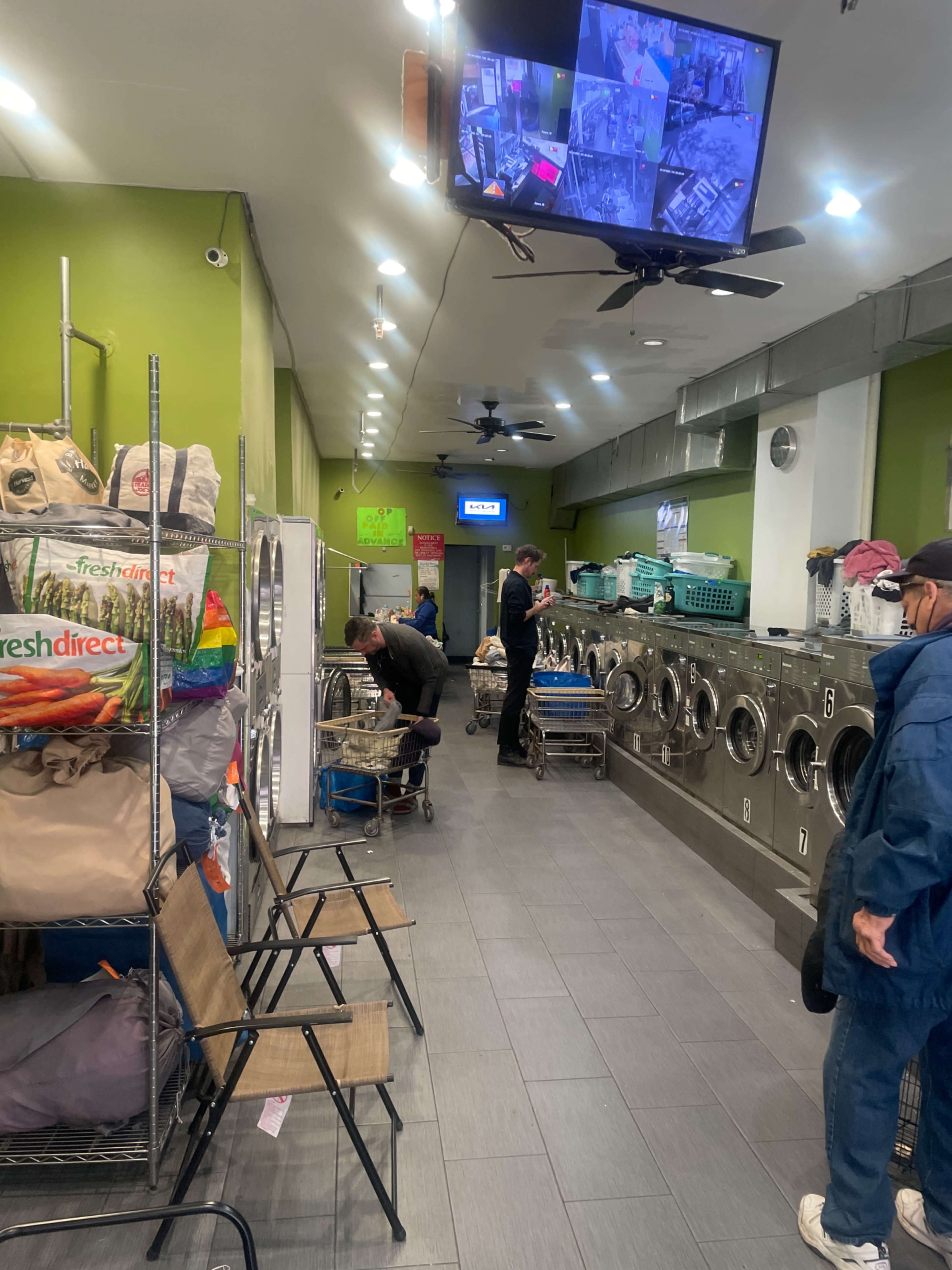 A laundromat with several washing machines, laundry carts, and patrons engaged in doing their laundry, while a security monitor displays camera footage.