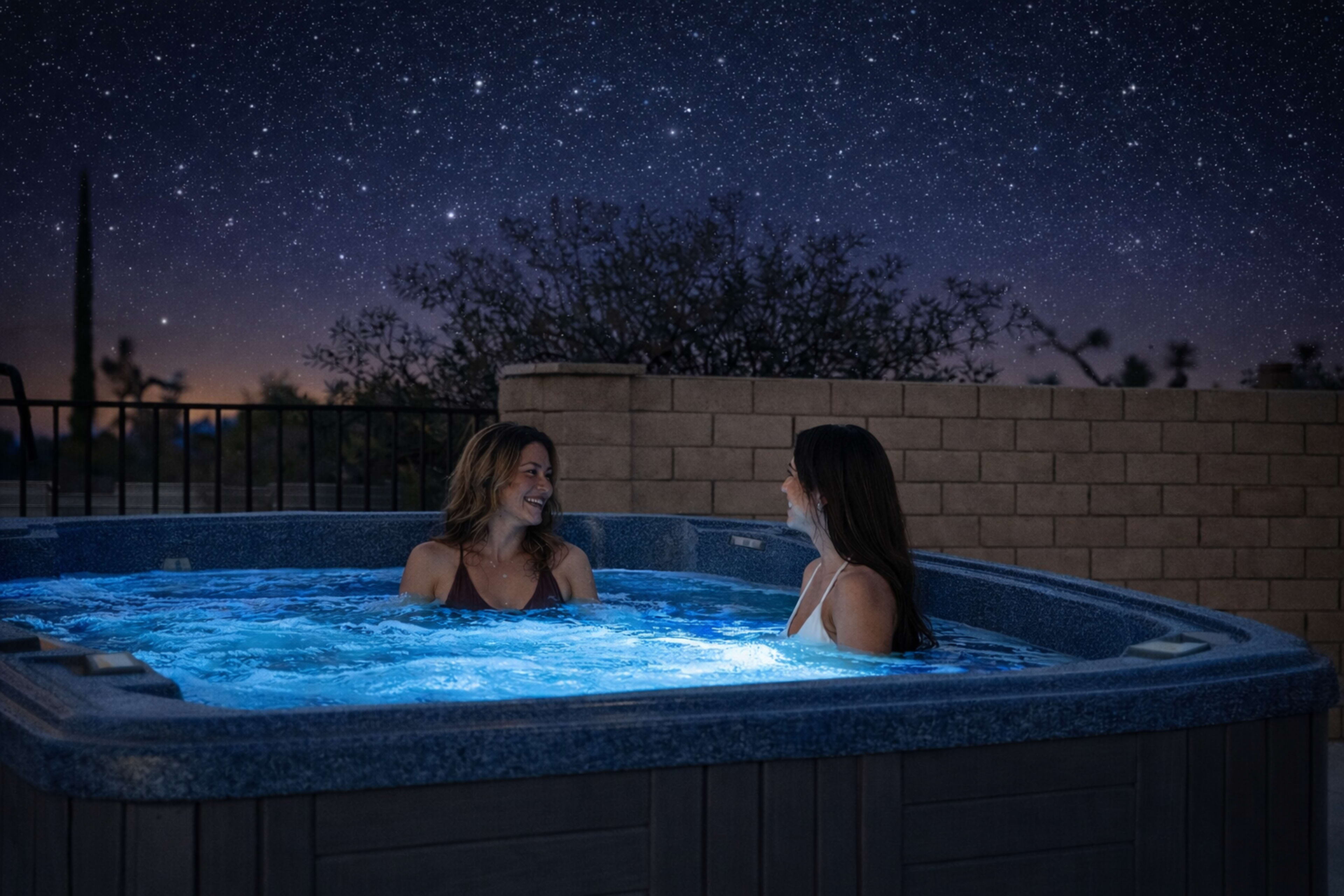 Two women are sitting in a hot tub under a starry night sky, with soft blue lights illuminating the water.