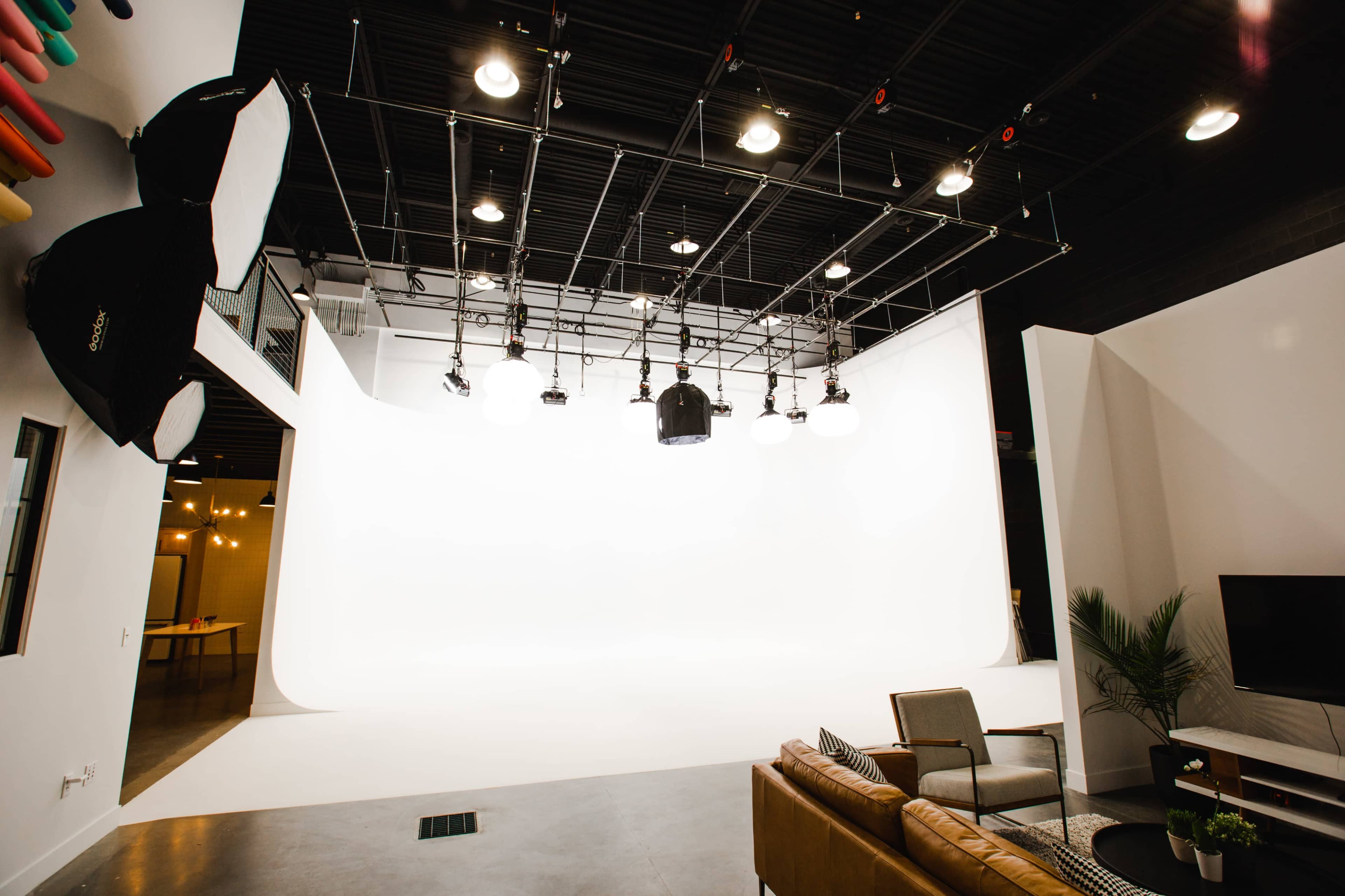 A modern studio space with a white backdrop, overhead lighting equipment, and a seating area with a couch and chairs.