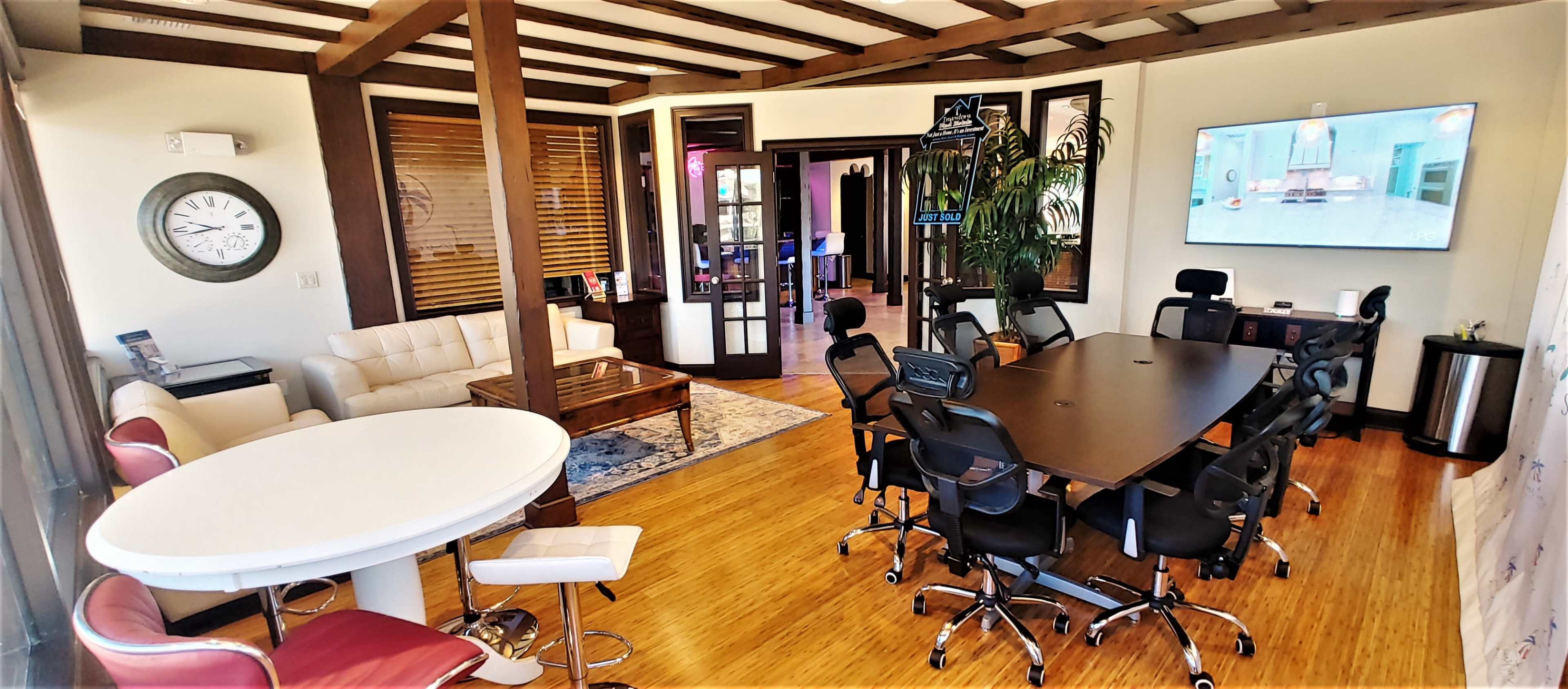 The image shows a modern office space featuring a large conference table surrounded by swivel chairs, a white round table with bar stools, a seating area with a couch, and a large screen on the wall.