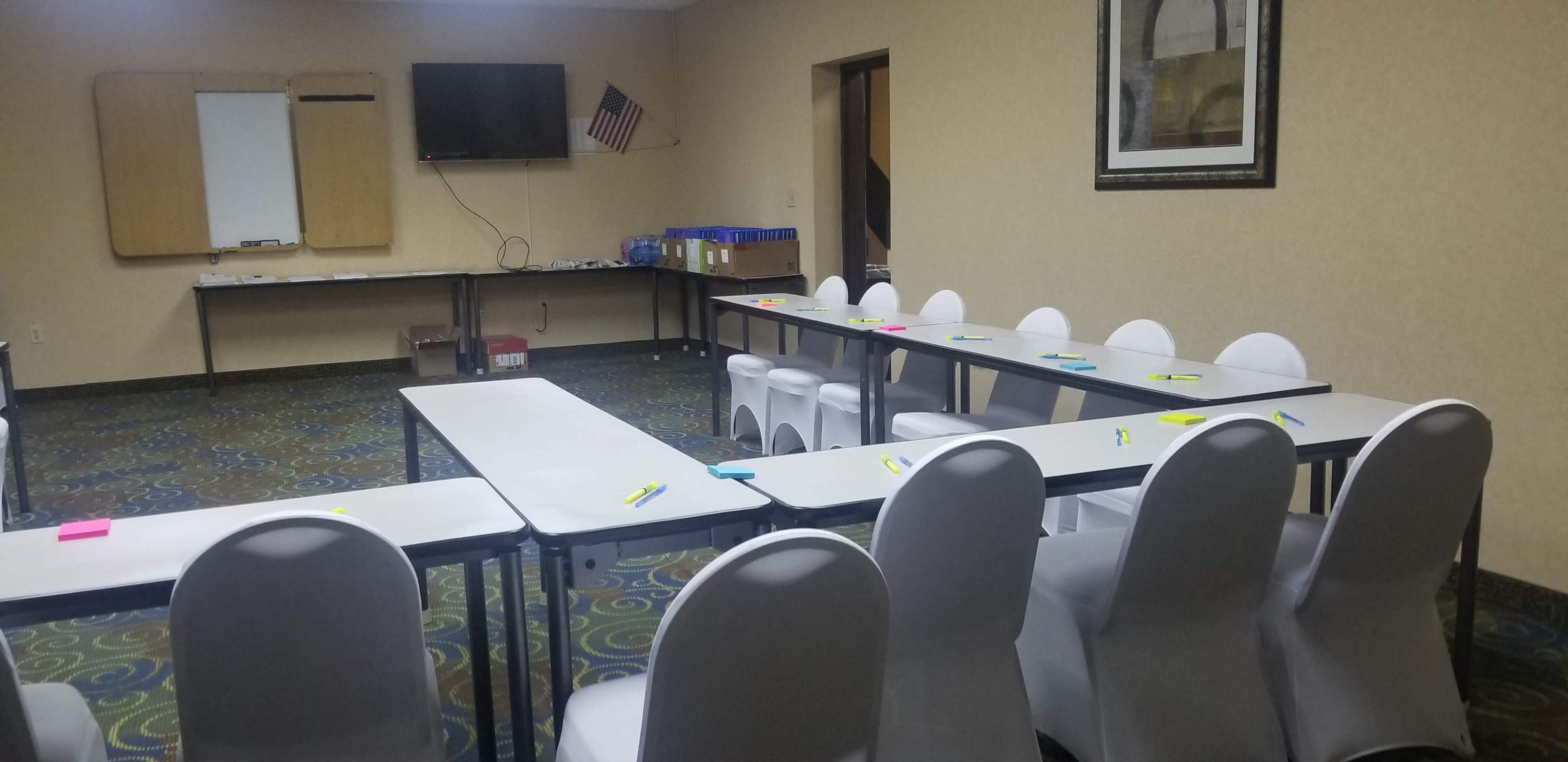 A classroom setting features several tables arranged in a U-shape, white chairs, and a television mounted on the wall, with an American flag in the background.