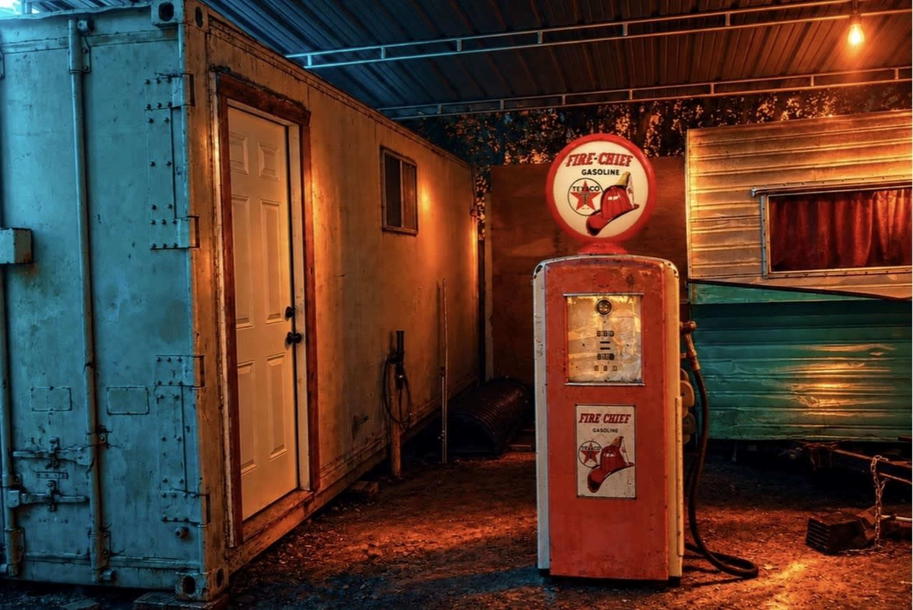 The image features a vintage gas pump in front of a trailer and a shipping container, illuminated by warm lighting.