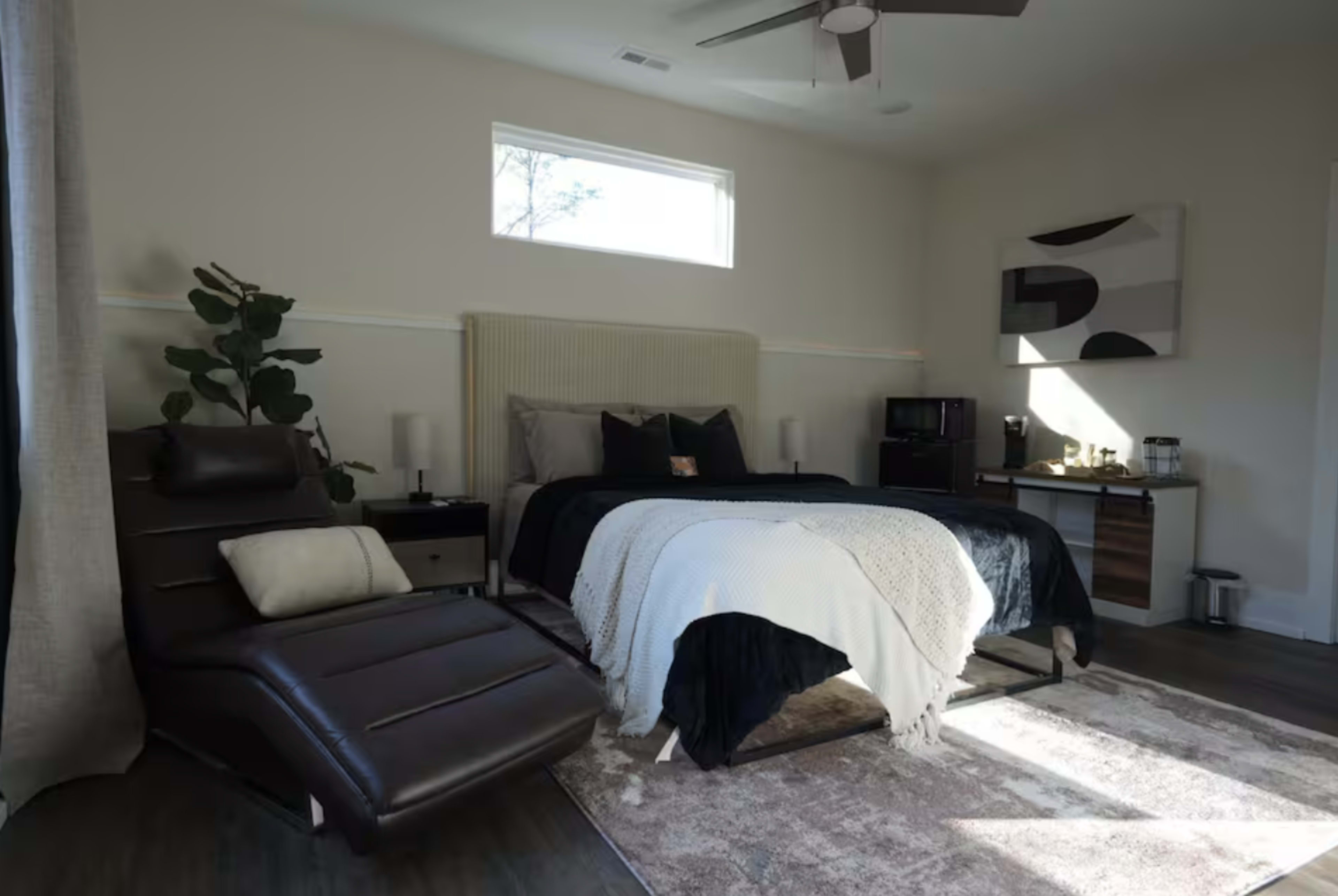 A modern bedroom features a large bed with dark bedding, a reclining chair, and a window letting in natural light.