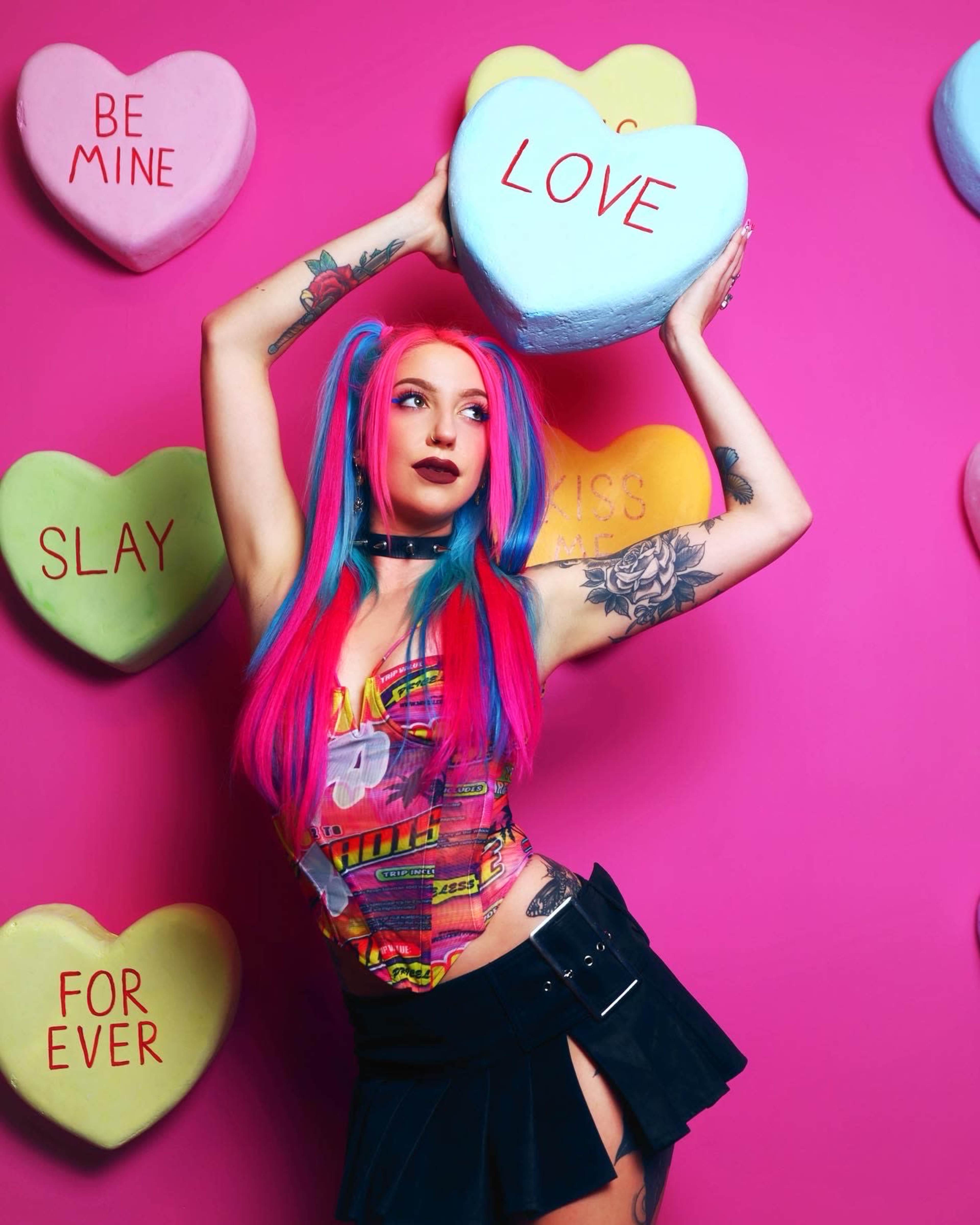 A woman with vibrant pink and blue hair holds large pastel-colored heart-shaped candies against a bright pink background.