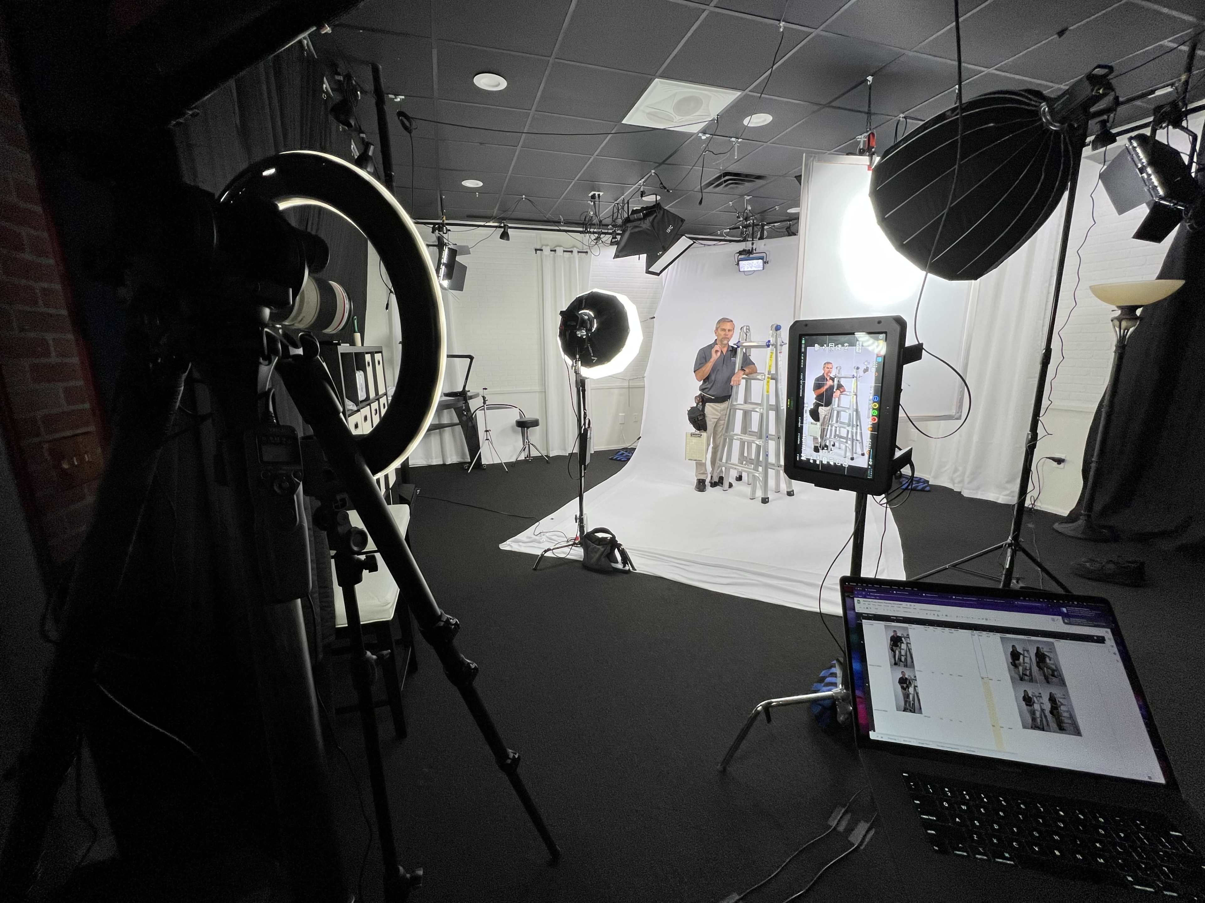 A photo studio setup features a seated subject under bright lights, with camera equipment, a laptop displaying images, and various lighting tools arranged around a white backdrop.