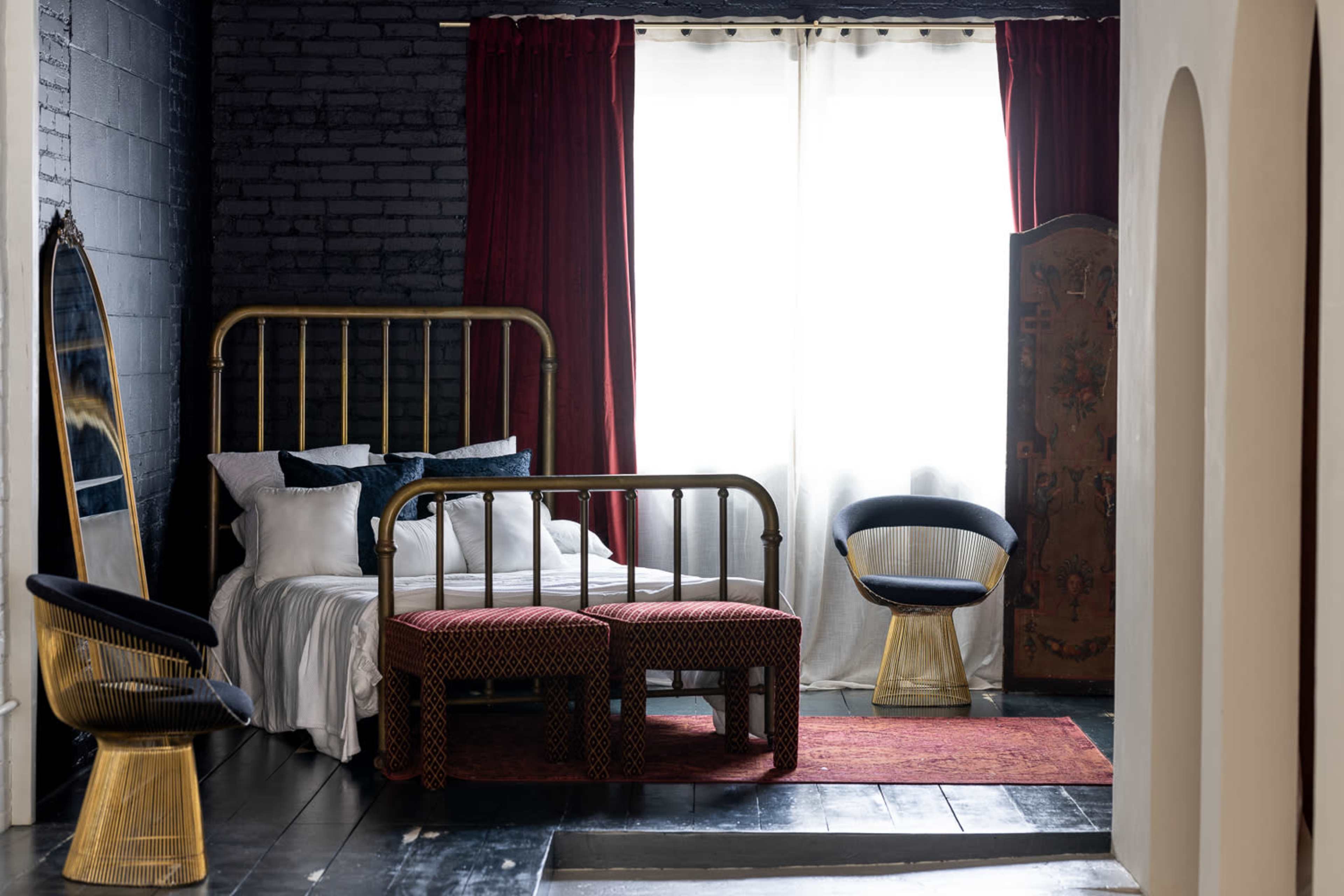 A bedroom with a brass bed frame, two upholstered stools, a red area rug, and large windows draped with white curtains.