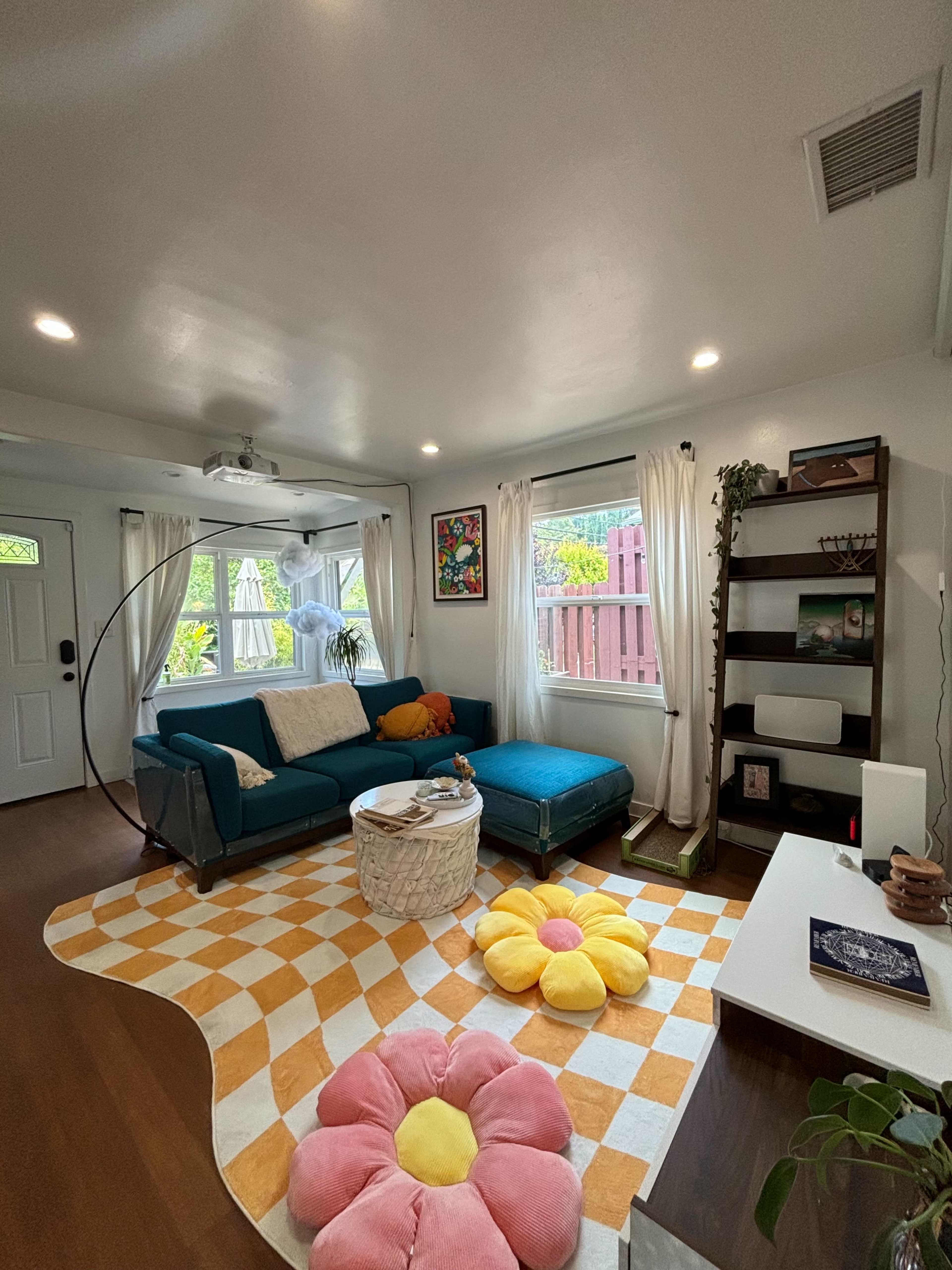 The room features a turquoise sofa, a round coffee table, a large pink flower-shaped pouf, and a bookshelf, all complemented by bright sunlight streaming through the windows.