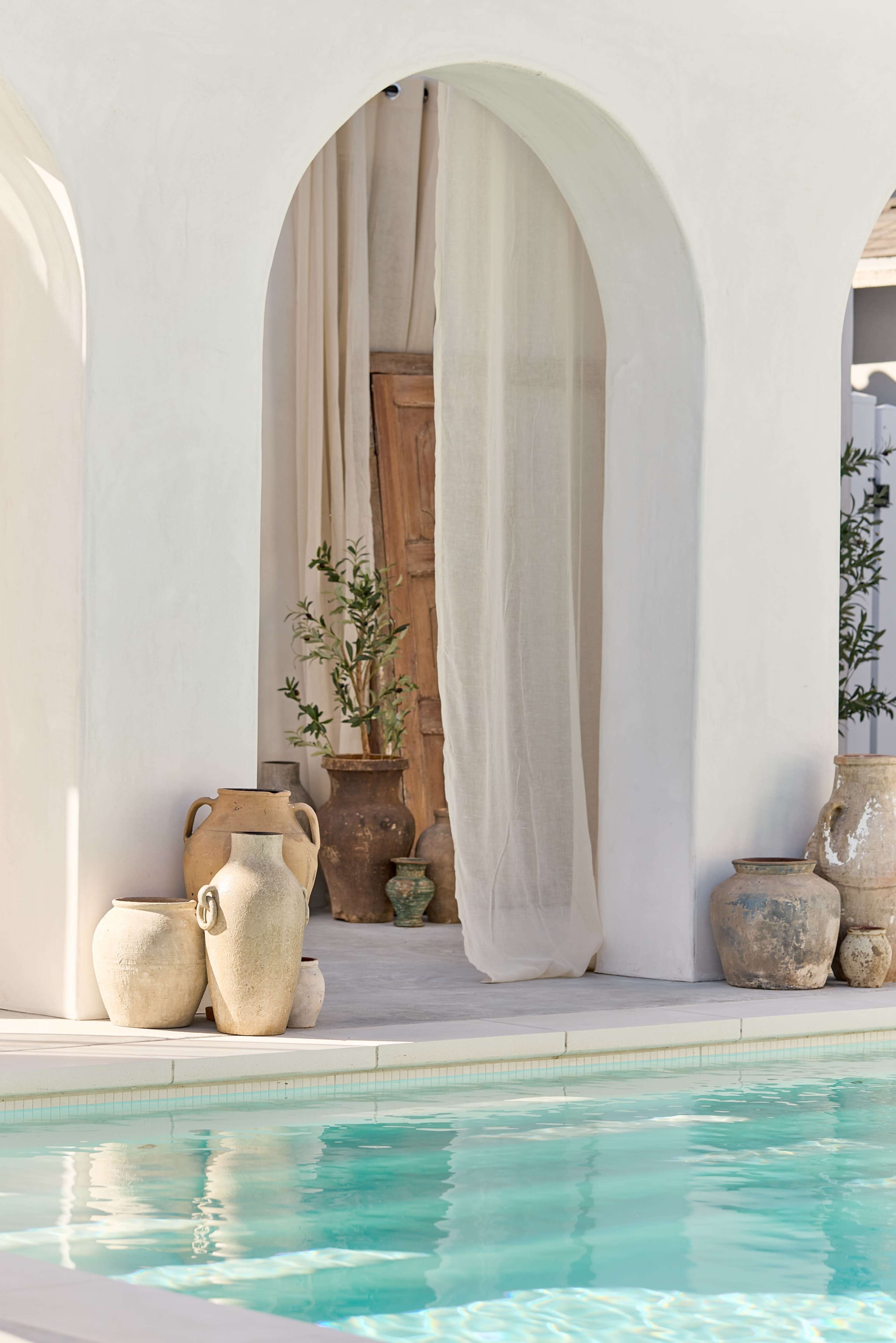 The image features a tranquil pool scene with decorative pottery arranged near an archway, complemented by sheer drapes and greenery in a light-colored architectural setting.
