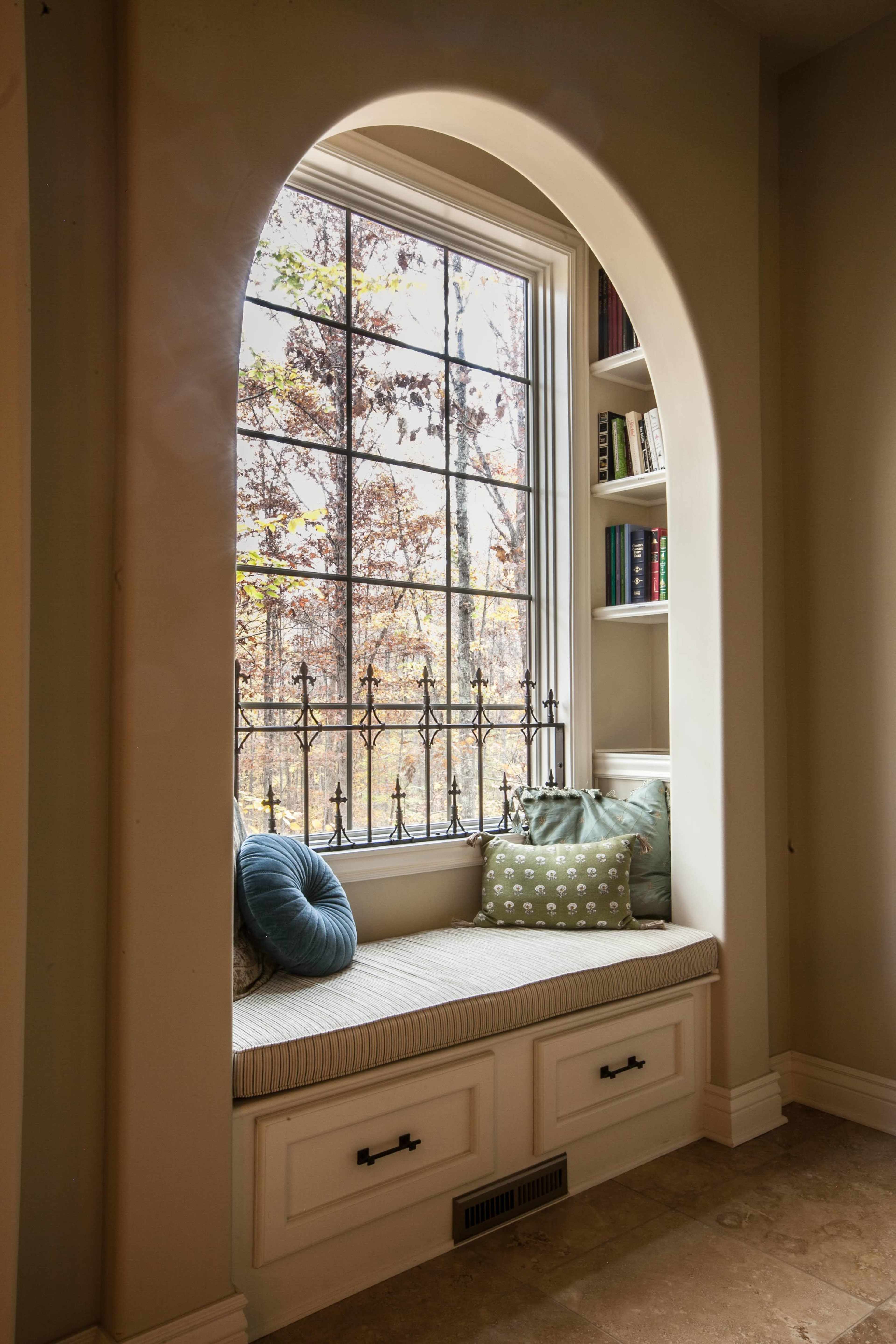 A cozy window seat with a cushion and pillows, framed by an arched opening and overlooking a wooded landscape through a large window.