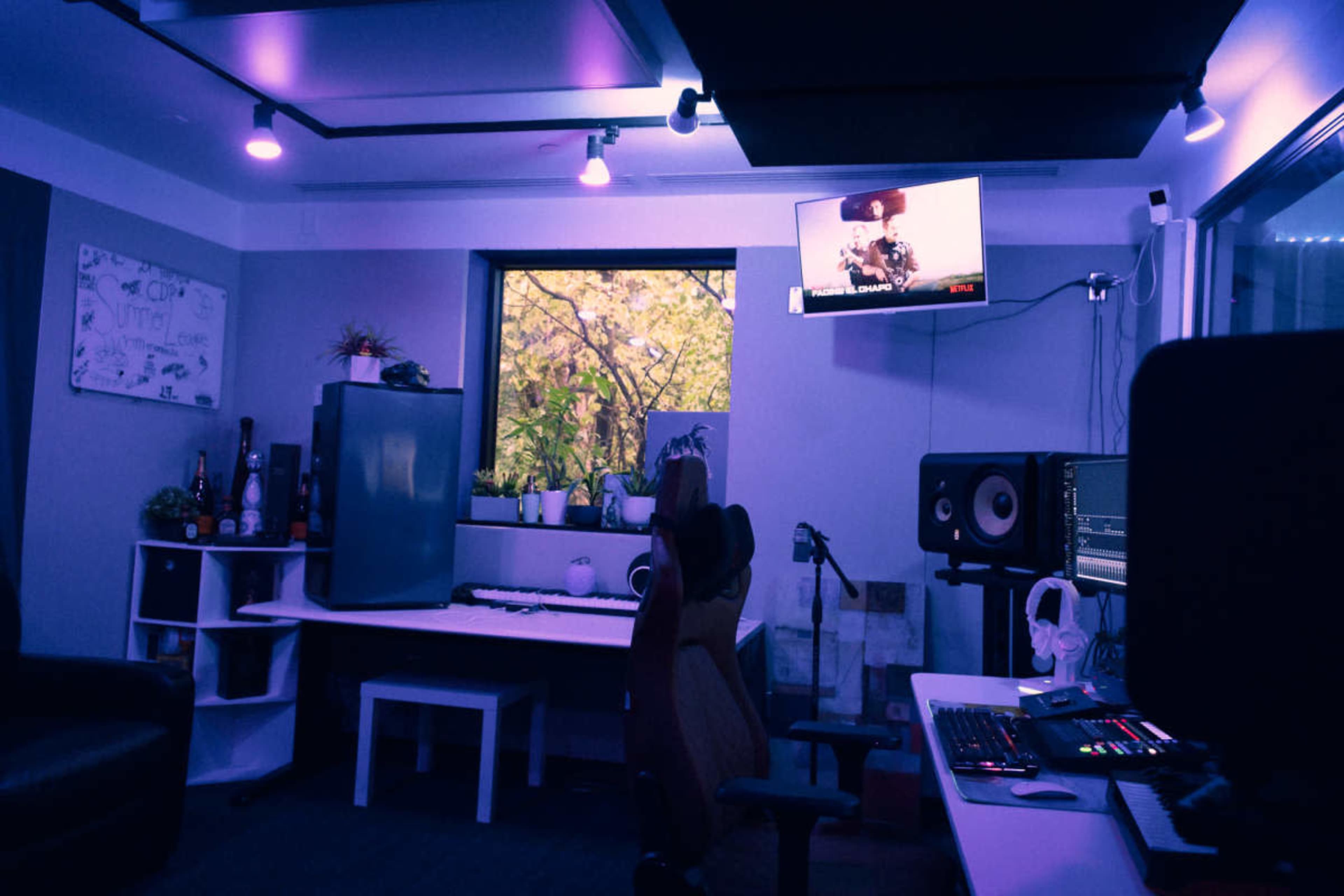 The image shows a dimly lit music studio featuring a desk with a keyboard, a computer screen displaying a video, and a window framed by autumn foliage.