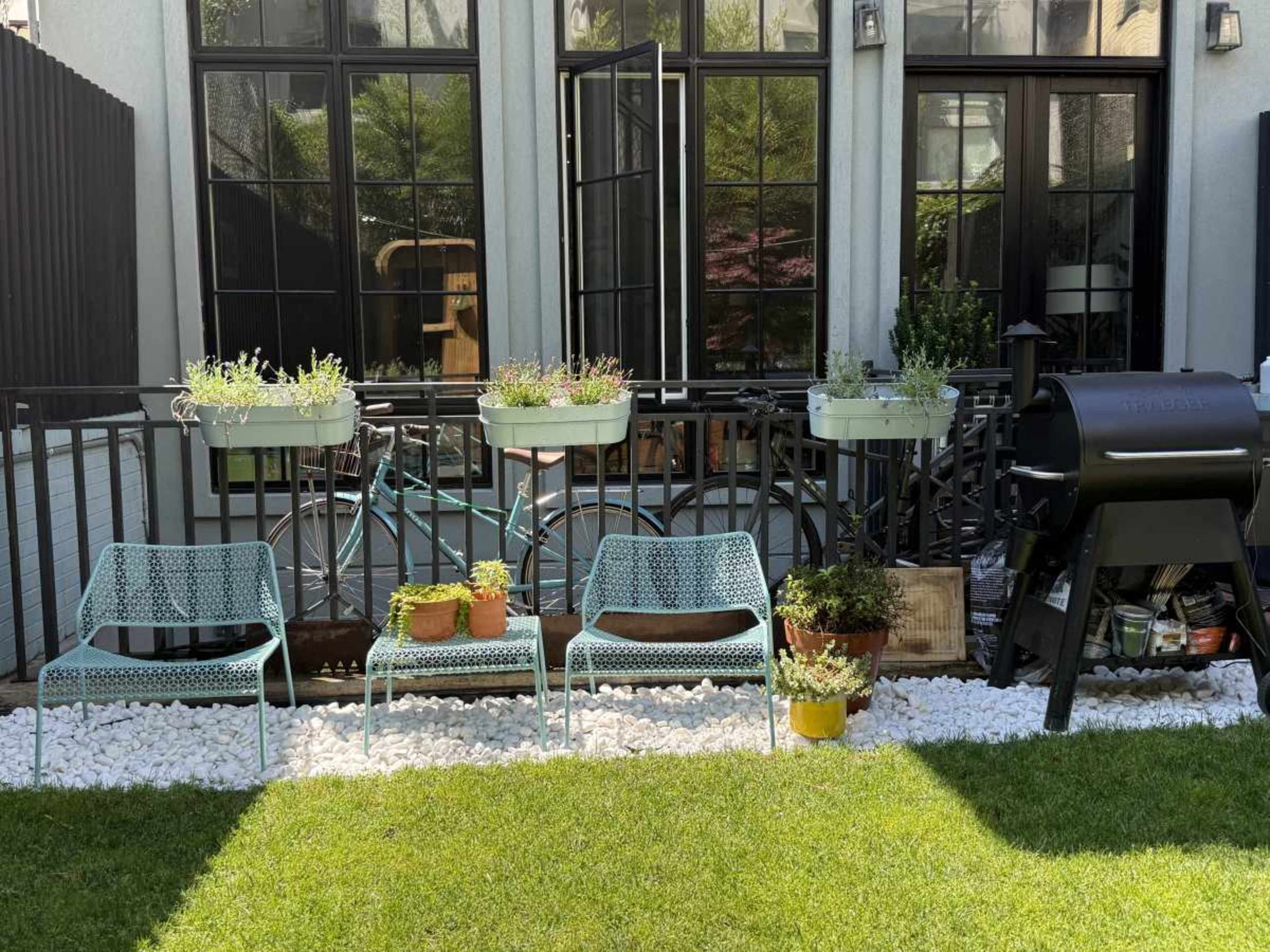 The image shows a small backyard patio with metal furniture, potted plants, and a barbecue grill, surrounded by a gravel area.