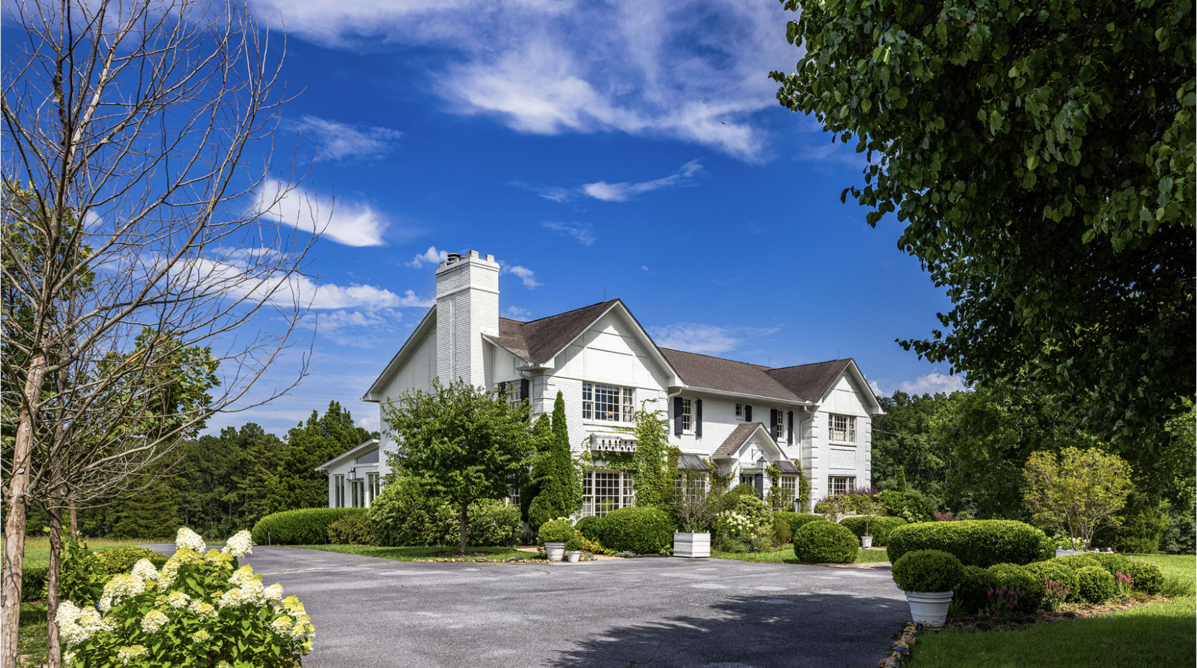 A large white house with a sloped roof is situated in a landscaped yard featuring trees and shrubs against a clear blue sky.