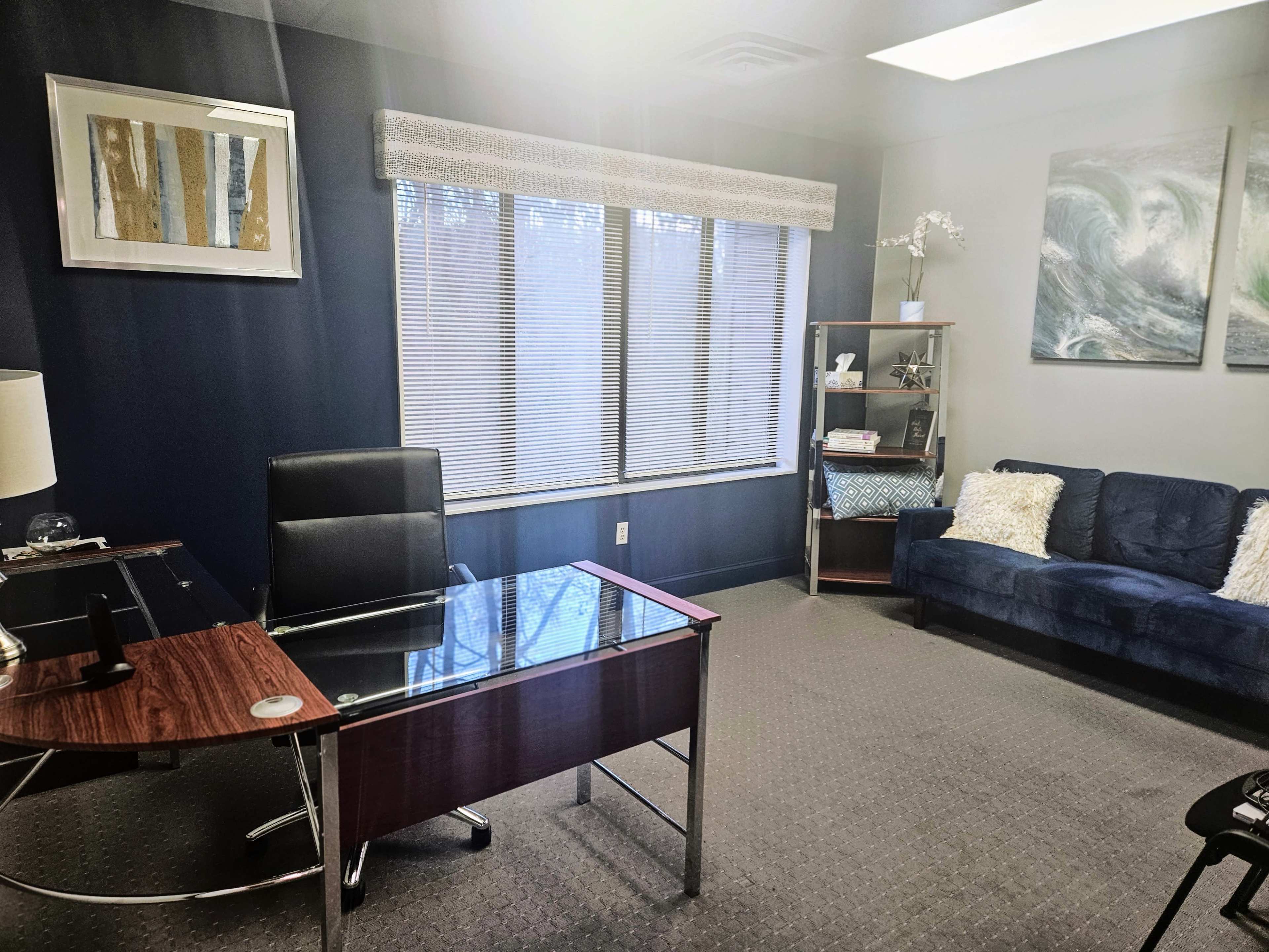 A spacious office featuring a wooden desk with a glass top, a black desk chair, a blue couch, and large windows adorned with blinds.