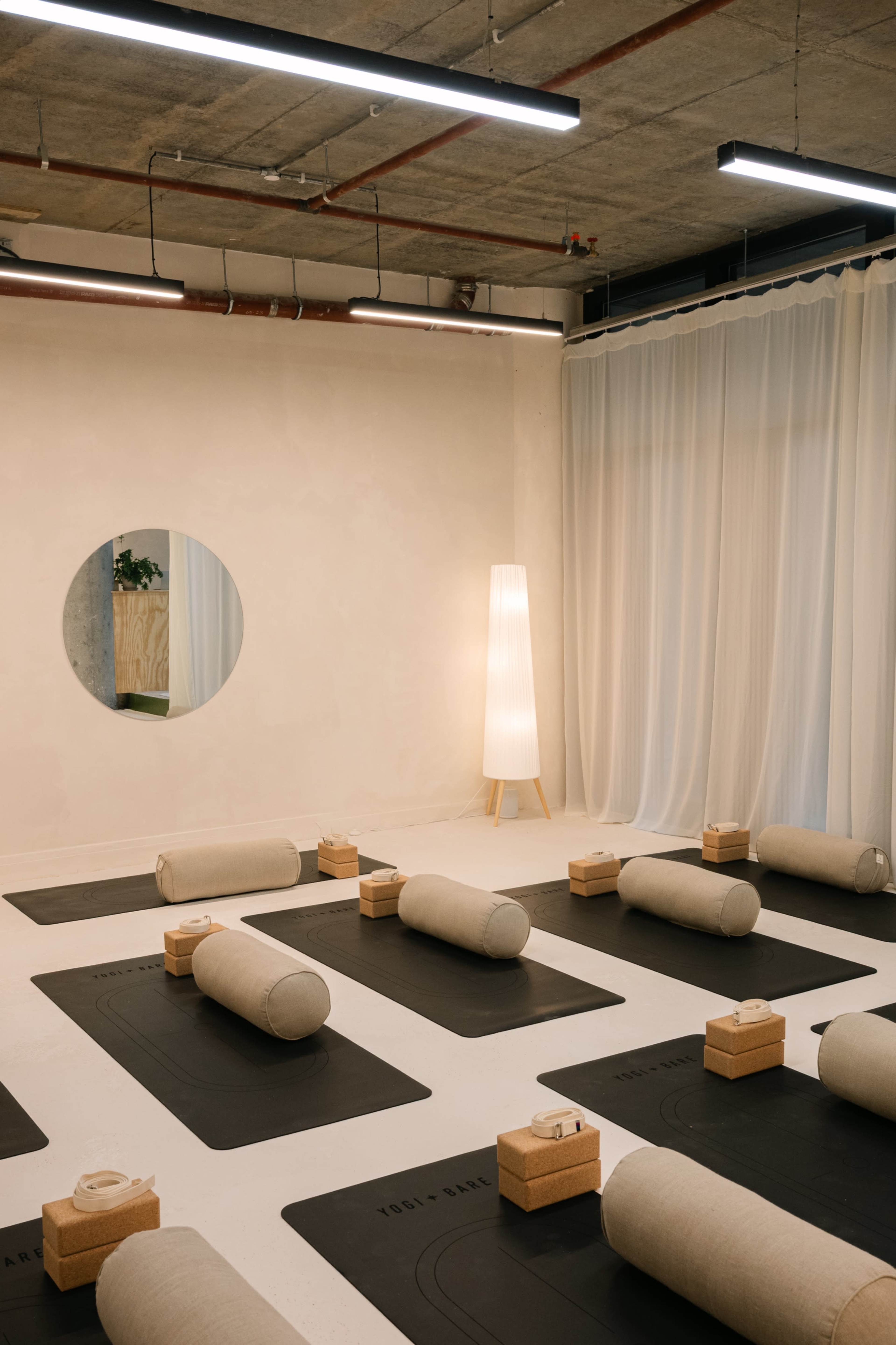 The image shows a minimalist yoga studio with cylindrical cushions on dark mats, wooden blocks, and a tall lamp in a softly lit space.