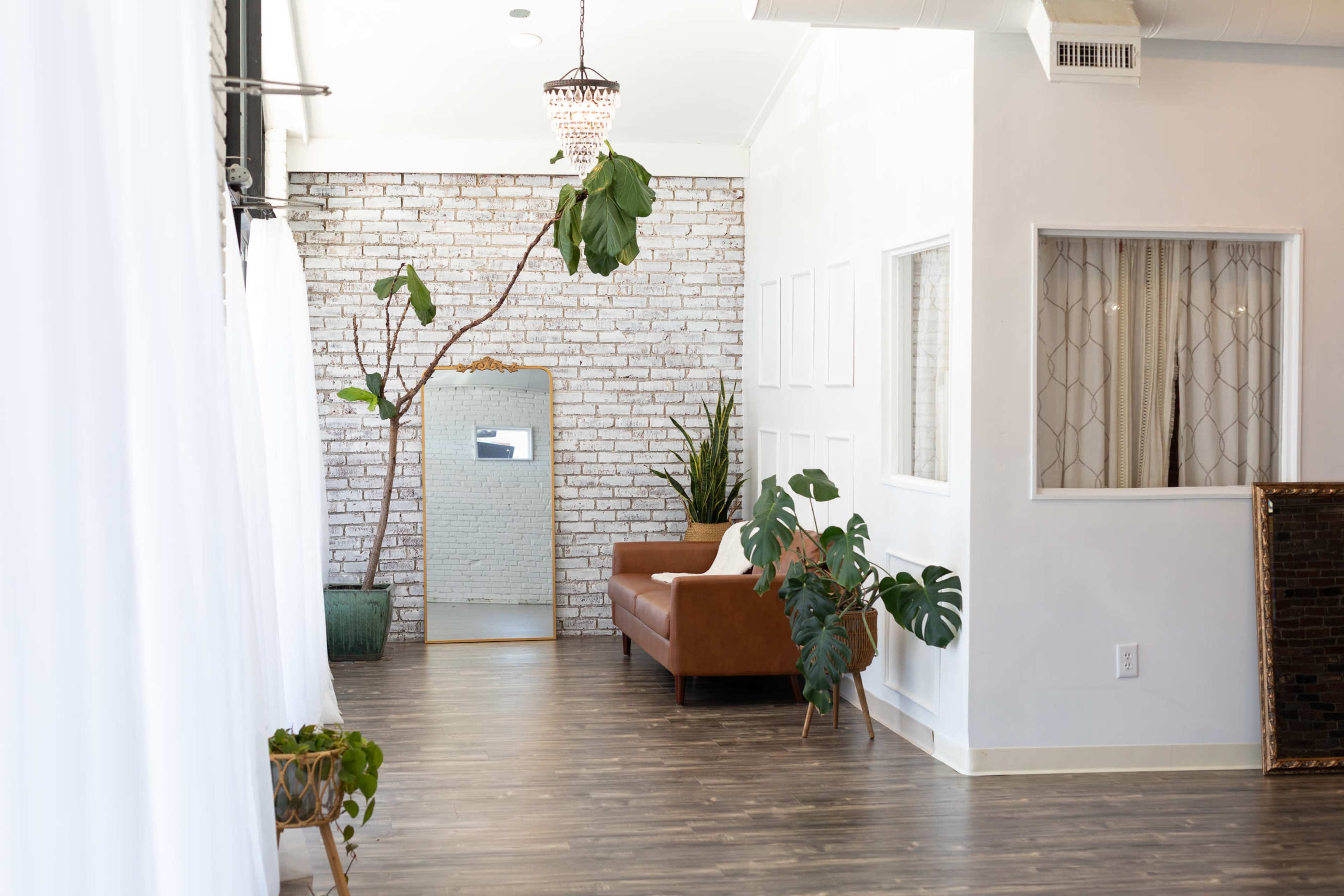 The image shows a bright, modern interior featuring a brown leather sofa, a large mirror, and decorative plants against a brick wall.