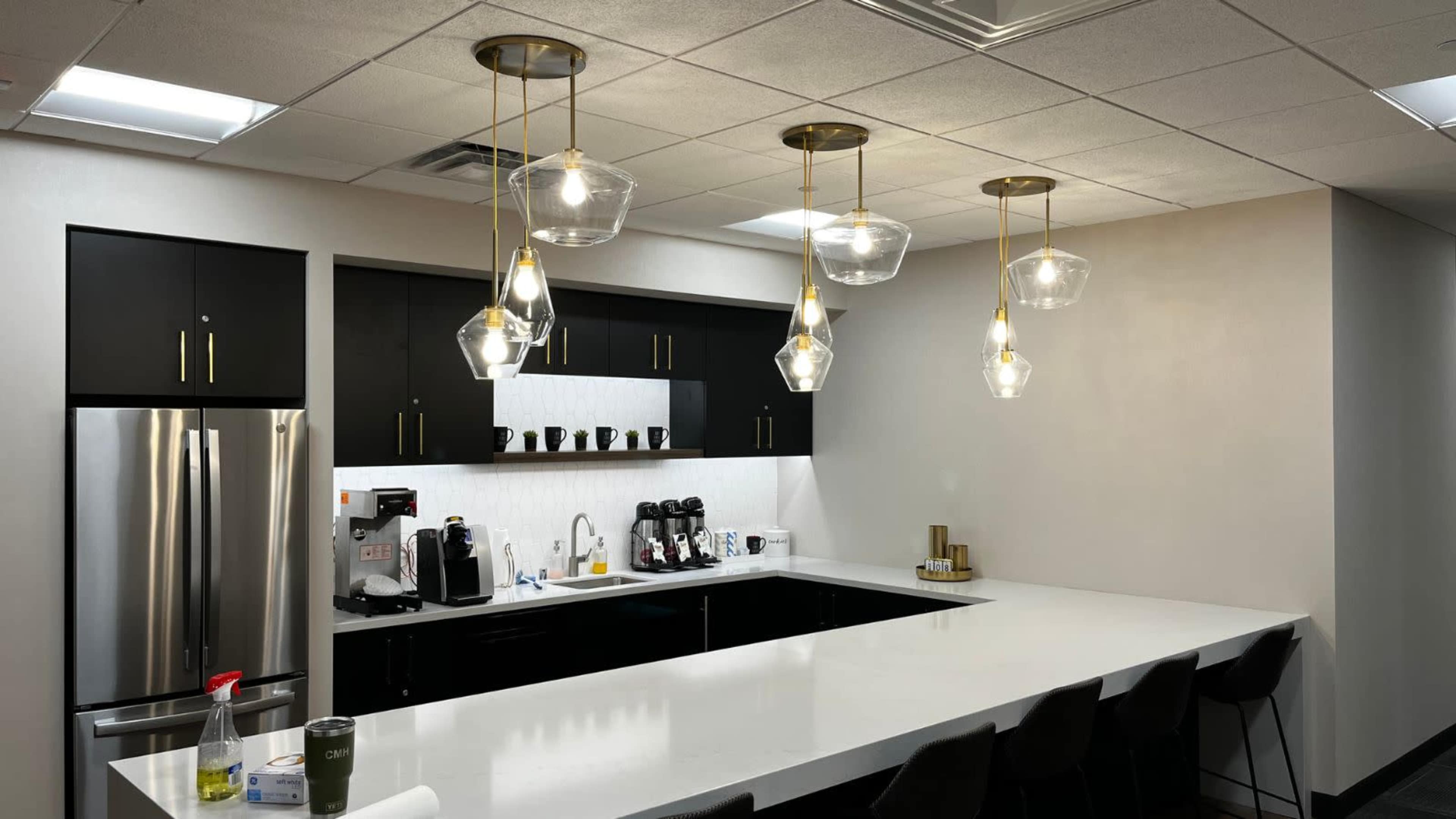 A modern kitchen with black cabinets, a long white countertop, stainless steel appliances, and pendant lighting.