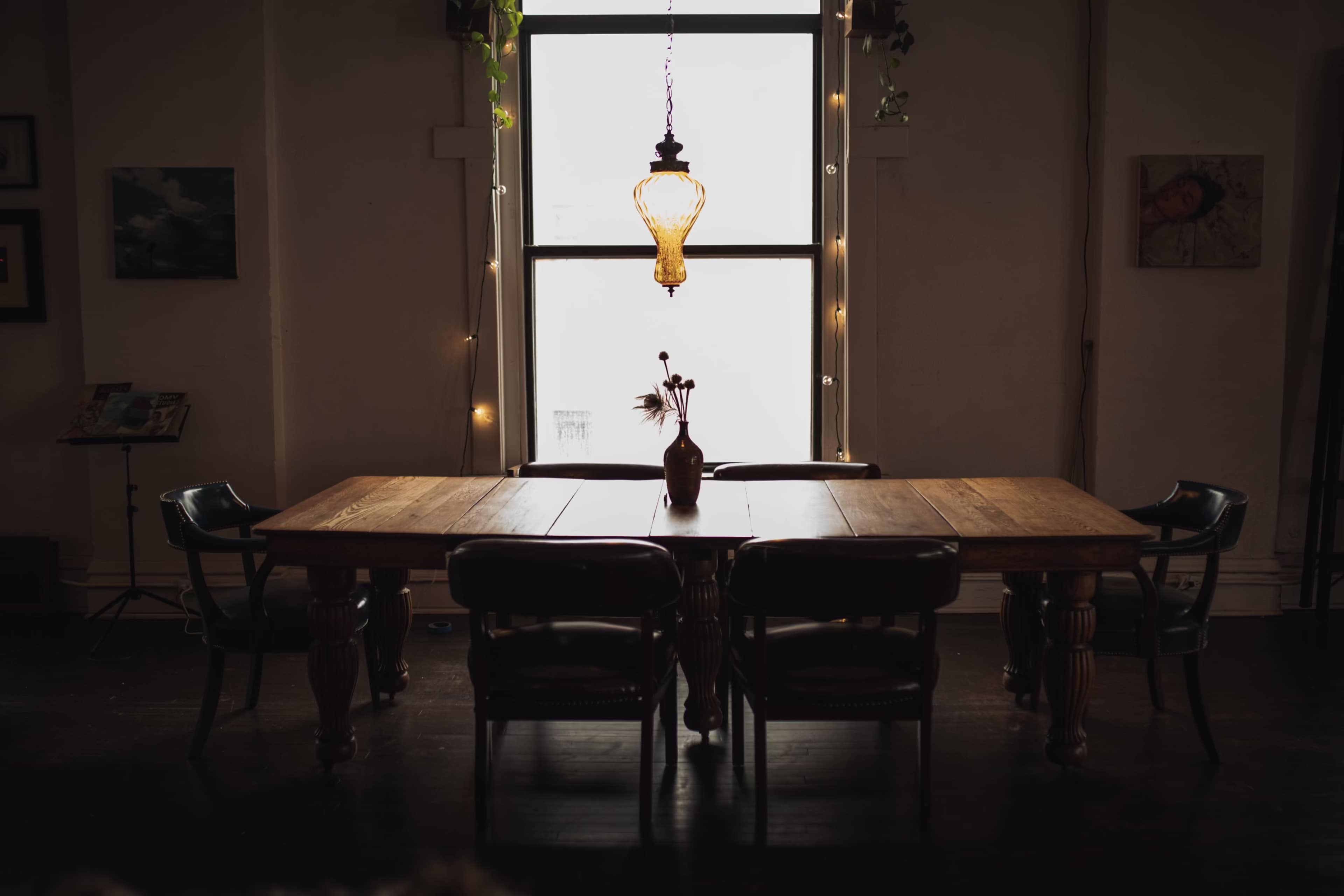 A wooden dining table surrounded by chairs is illuminated by a hanging light fixture in a dimly lit room.