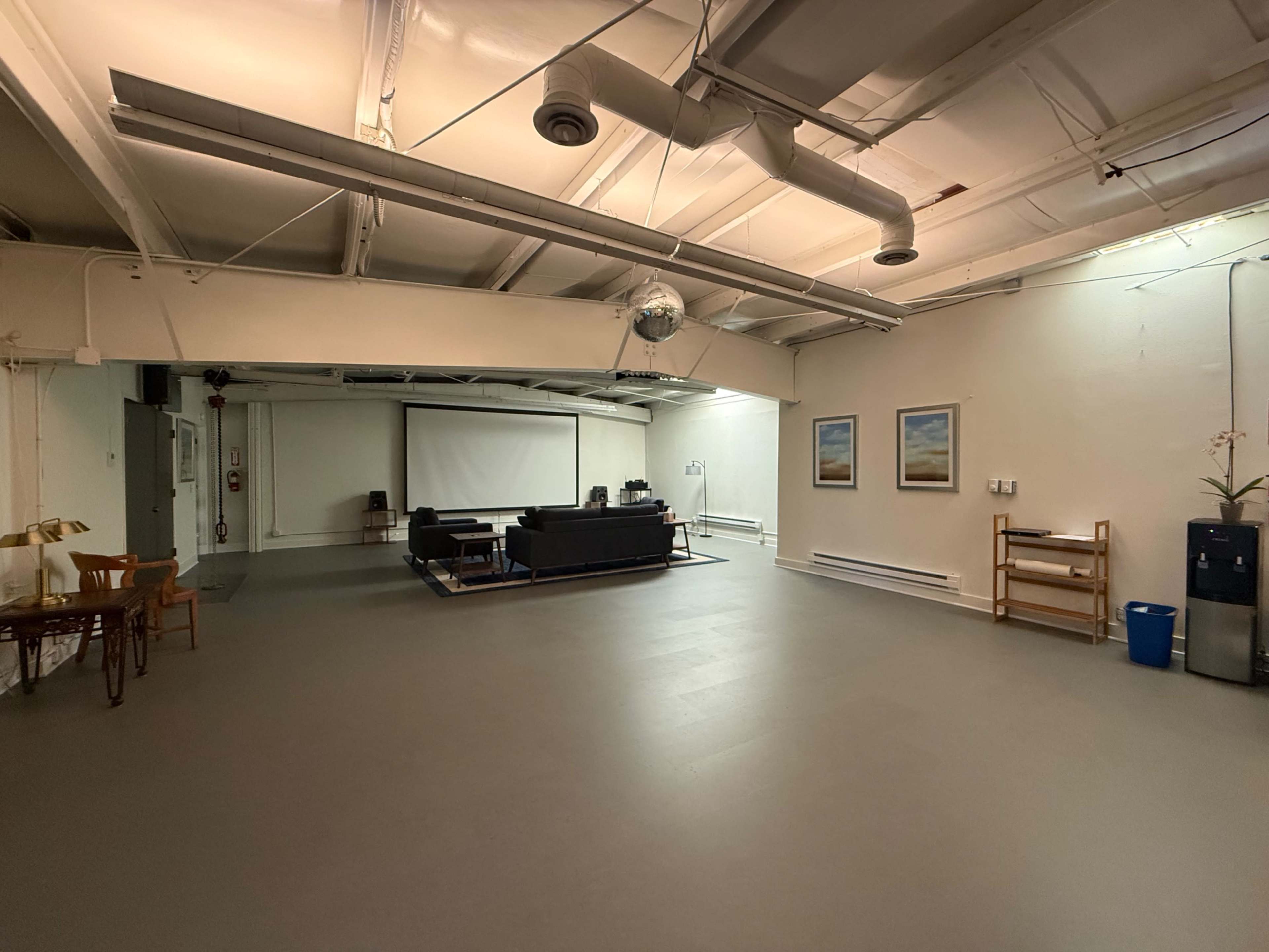 The image shows a spacious, minimalist indoor area featuring a sofa set, a projector screen, two framed pictures on the wall, and some furniture arranged along the sides.