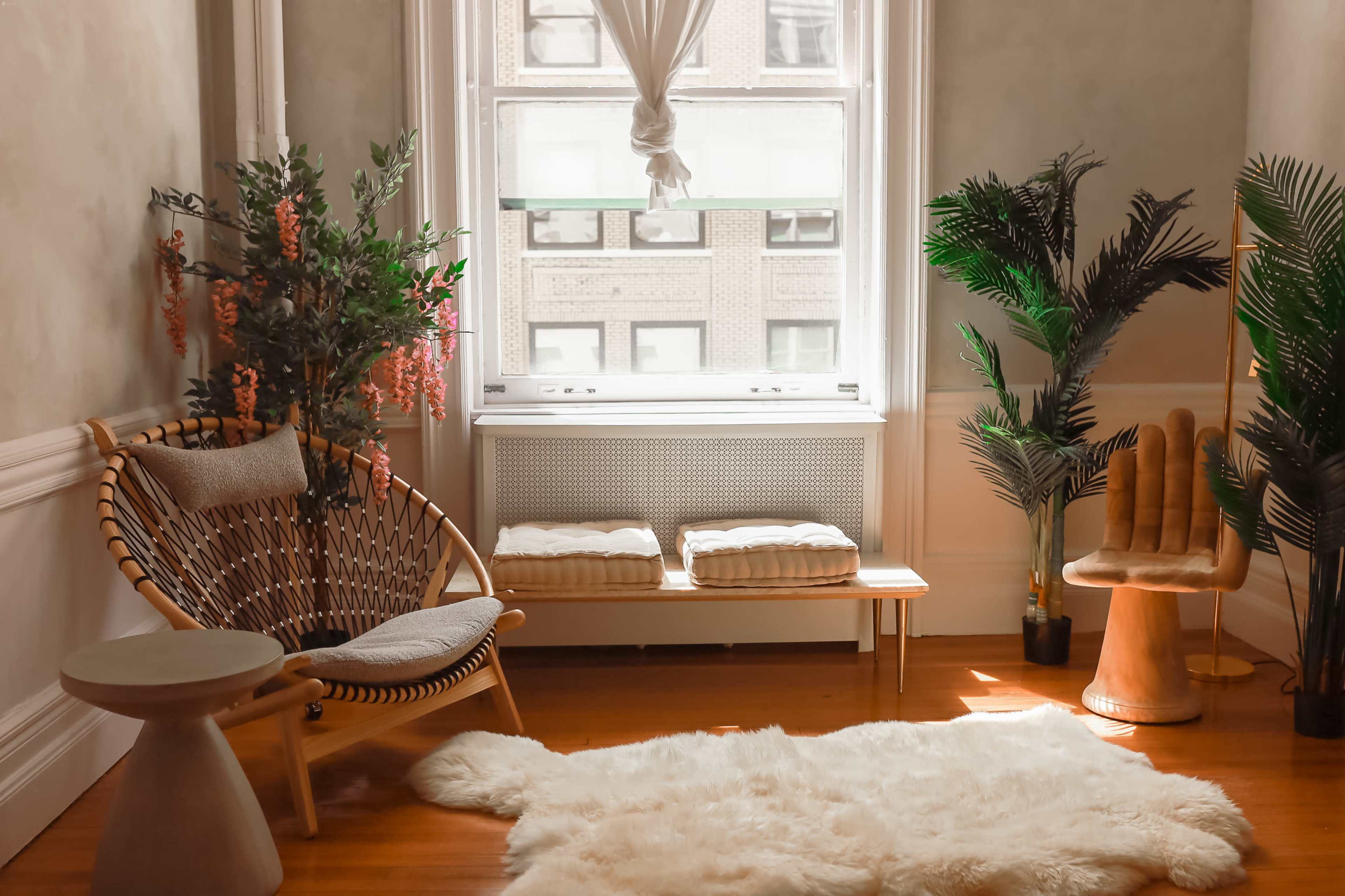 A cozy corner features a rattan chair, a small table, a bench, and lush greenery by a window.