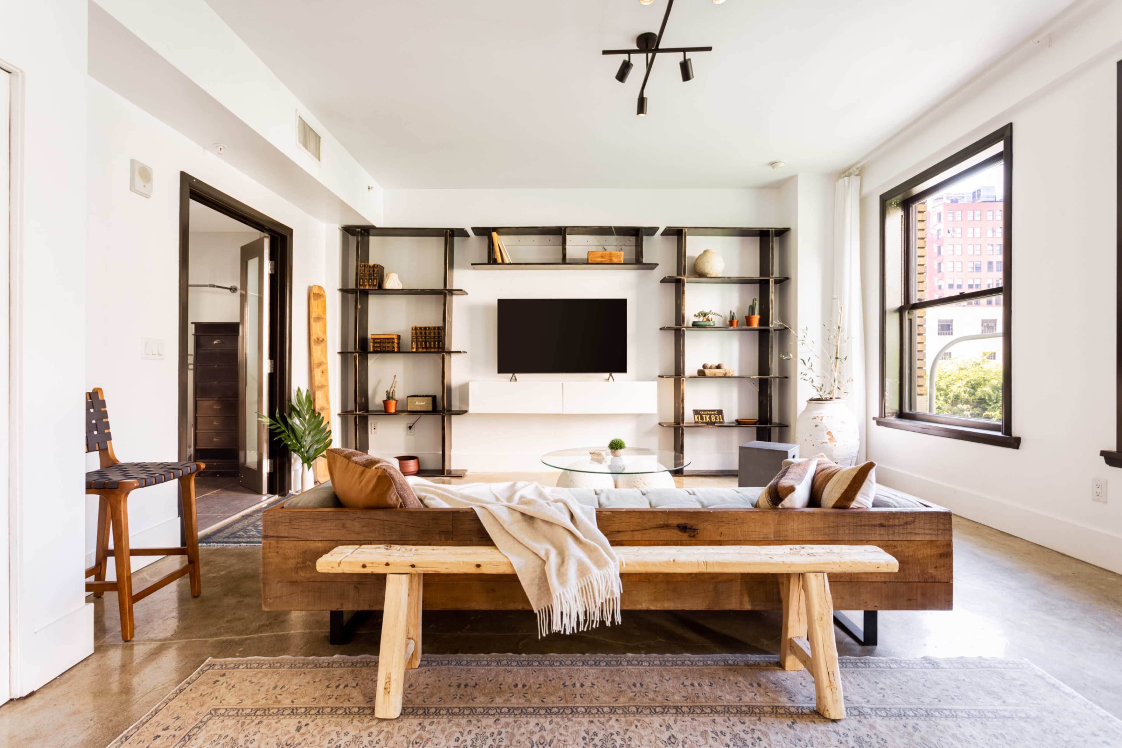 A modern living room with a wooden sofa, a glass coffee table, a flat-screen TV mounted on a wall, and shelves filled with decorative items.