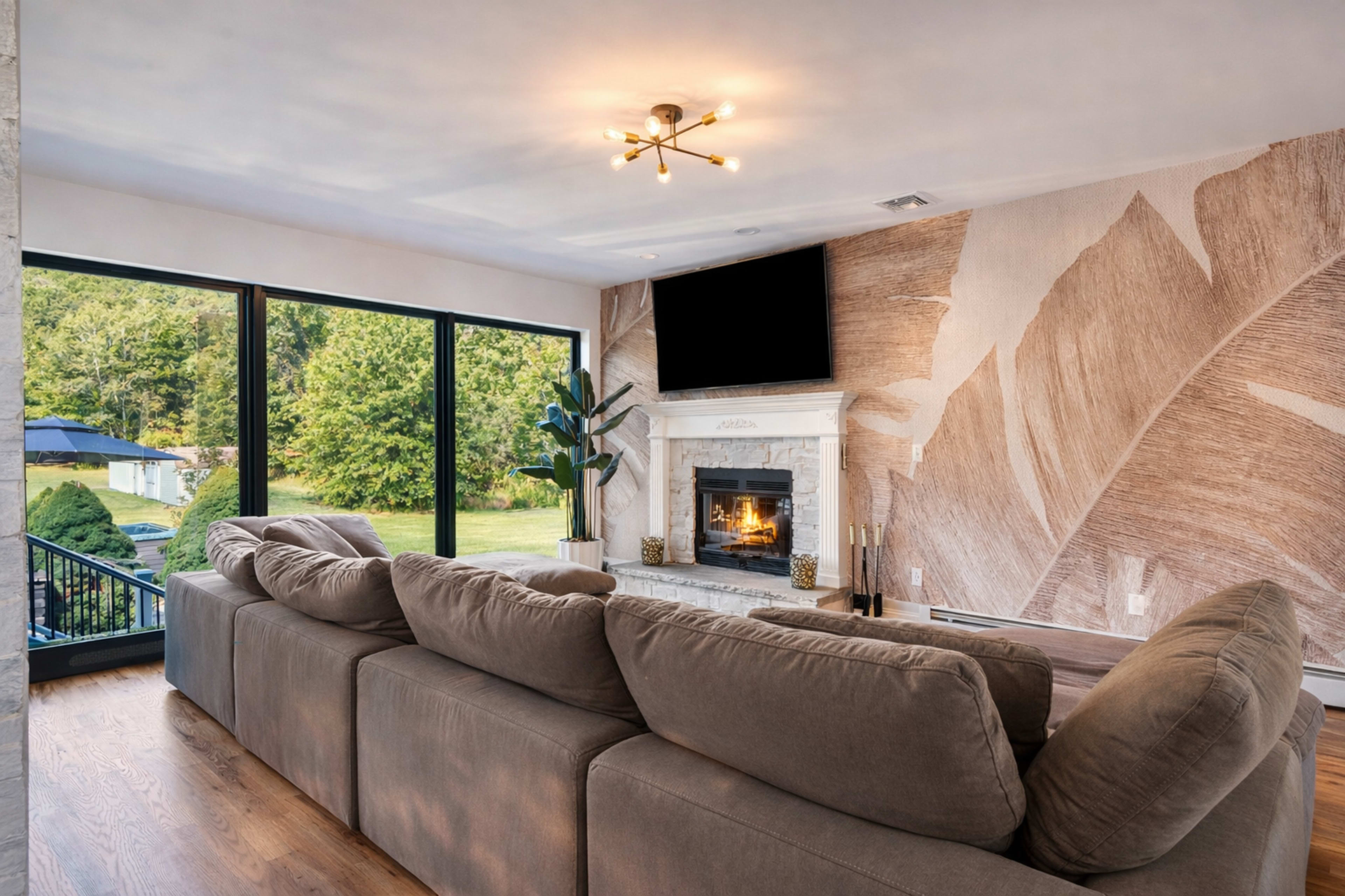 A modern living room features a large sectional sofa facing a fireplace, with large windows that provide a view of the outdoors.