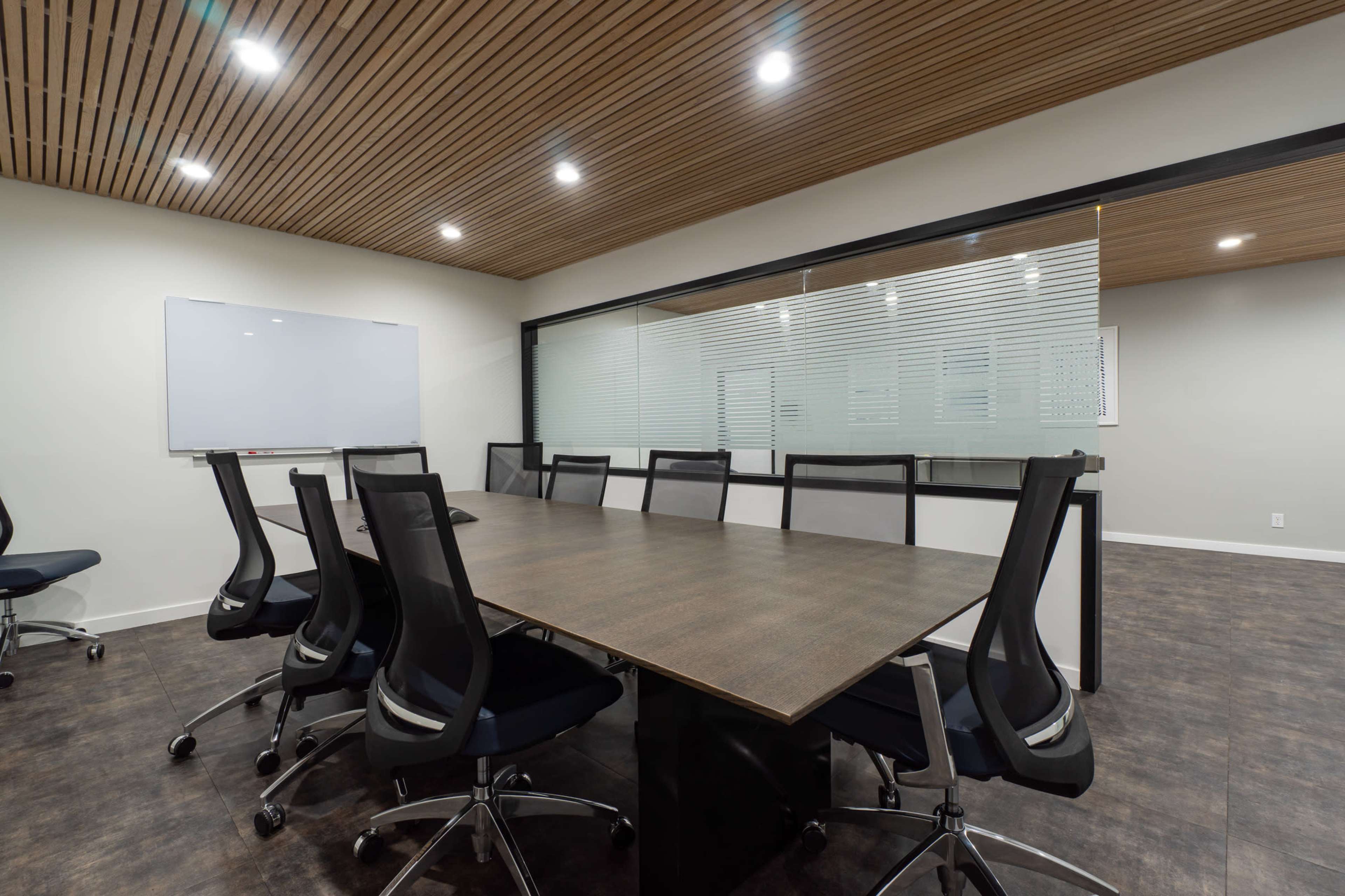 A conference room features a long wooden table surrounded by black ergonomic chairs, with a whiteboard and glass partition in the background.