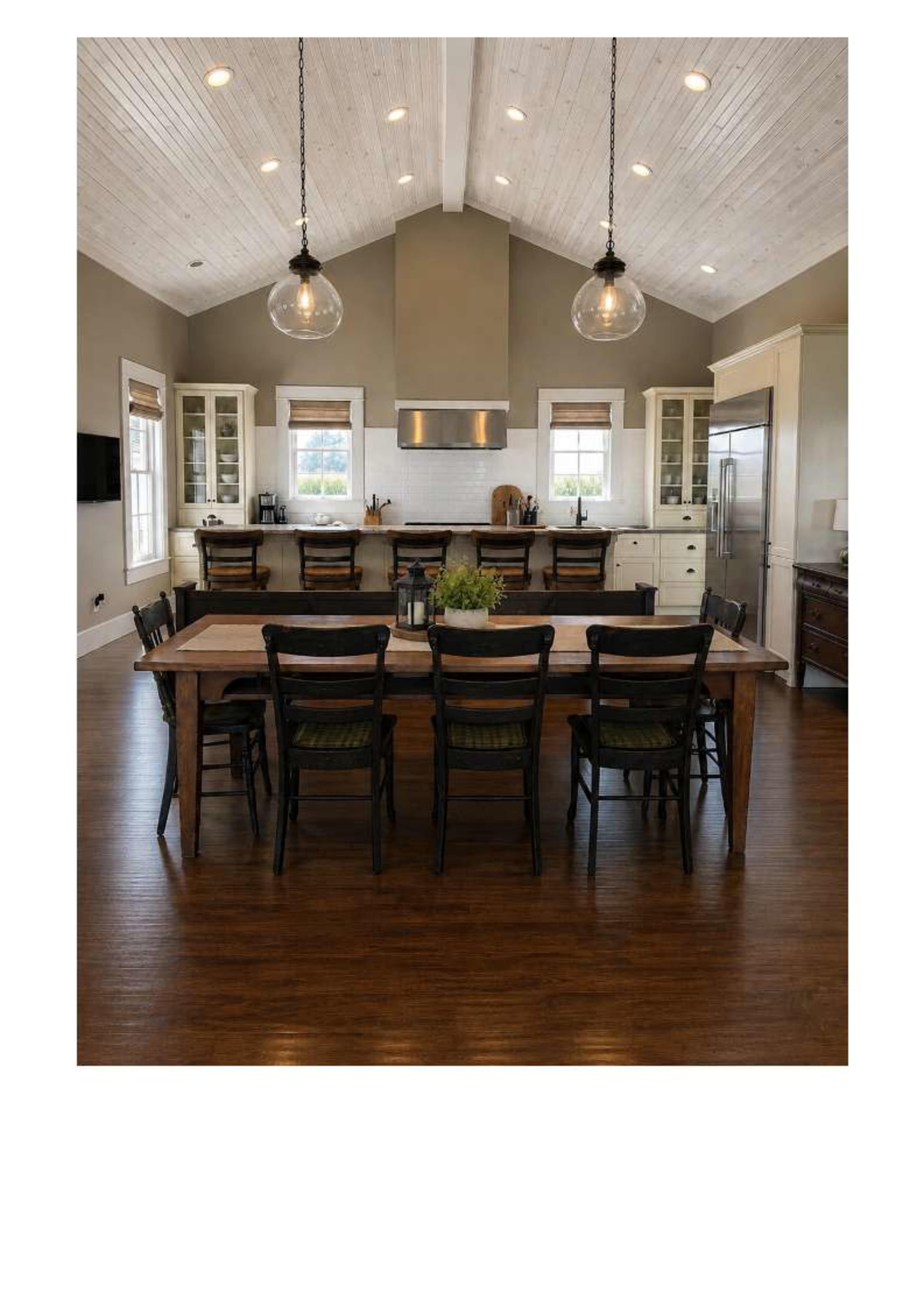 A spacious kitchen features a long wooden dining table surrounded by black chairs, with large windows and a high ceiling illuminated by pendant lights.
