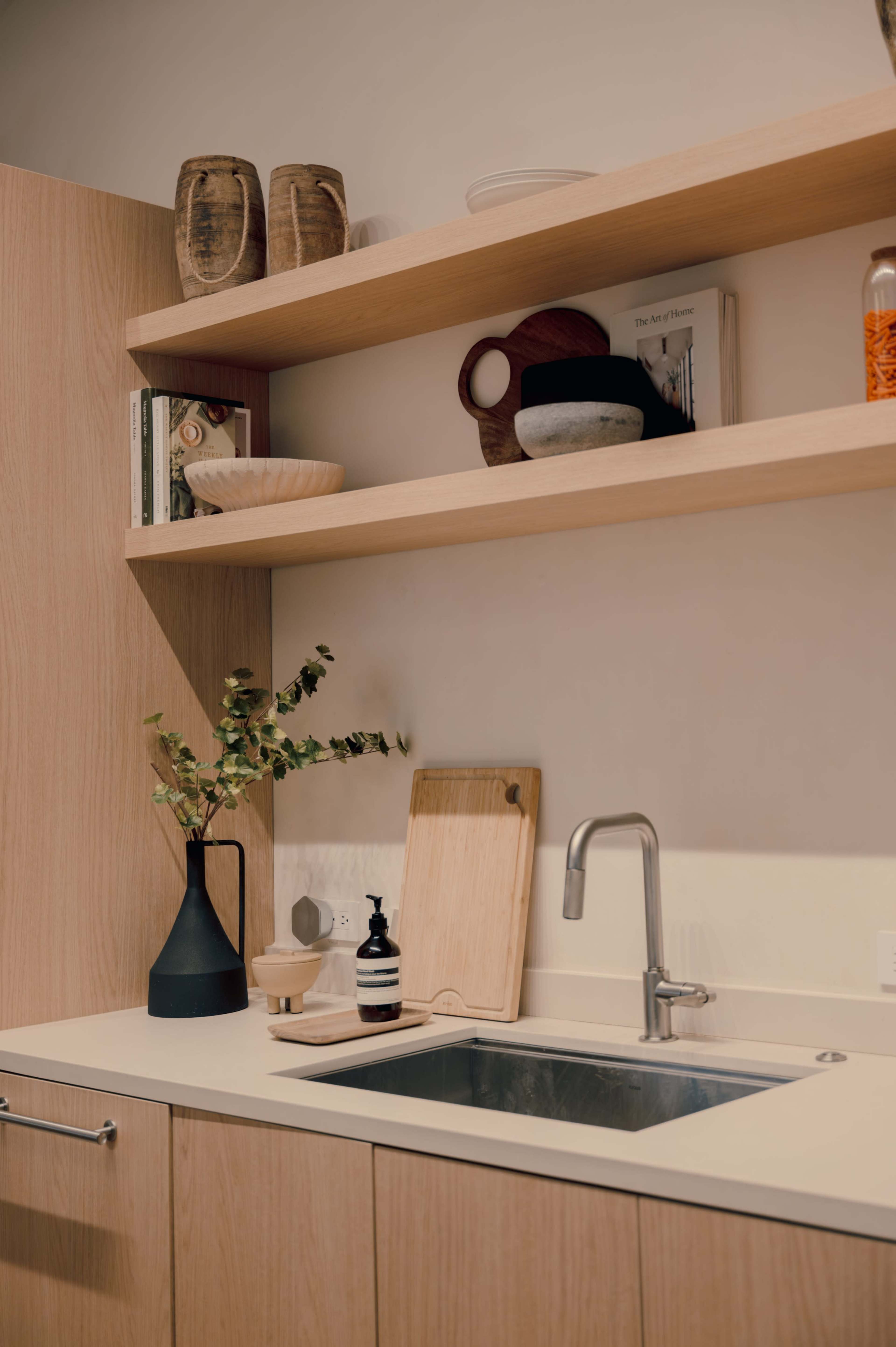 A modern kitchen features wooden shelves with various kitchen items, a plant in a vase, and a sink with a cutting board and soap dispenser nearby.