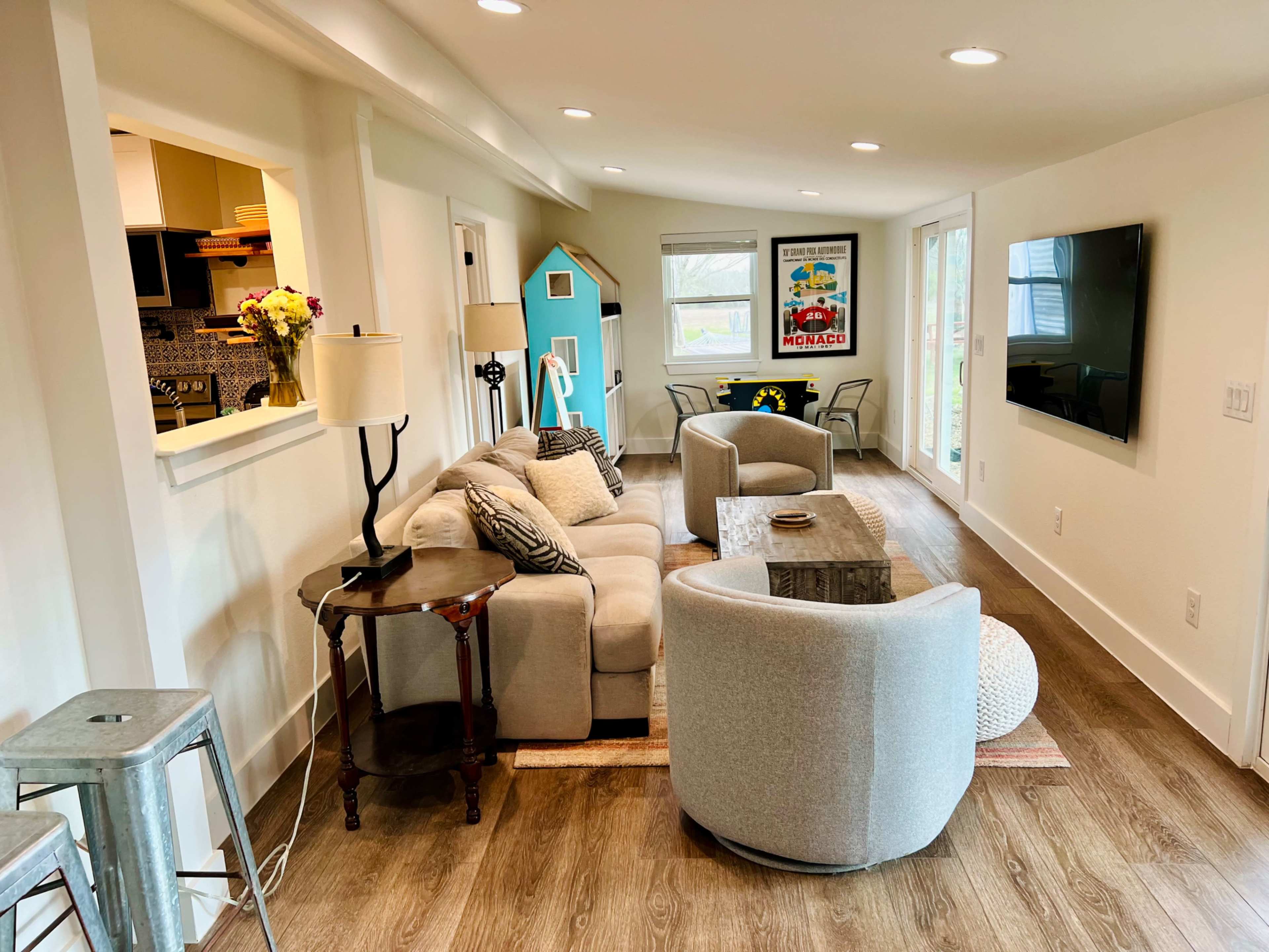 The image shows a modern, well-furnished living room with a sofa, two armchairs, a coffee table, and a television mounted on the wall, alongside large windows and a decorative blue house plant stand.
