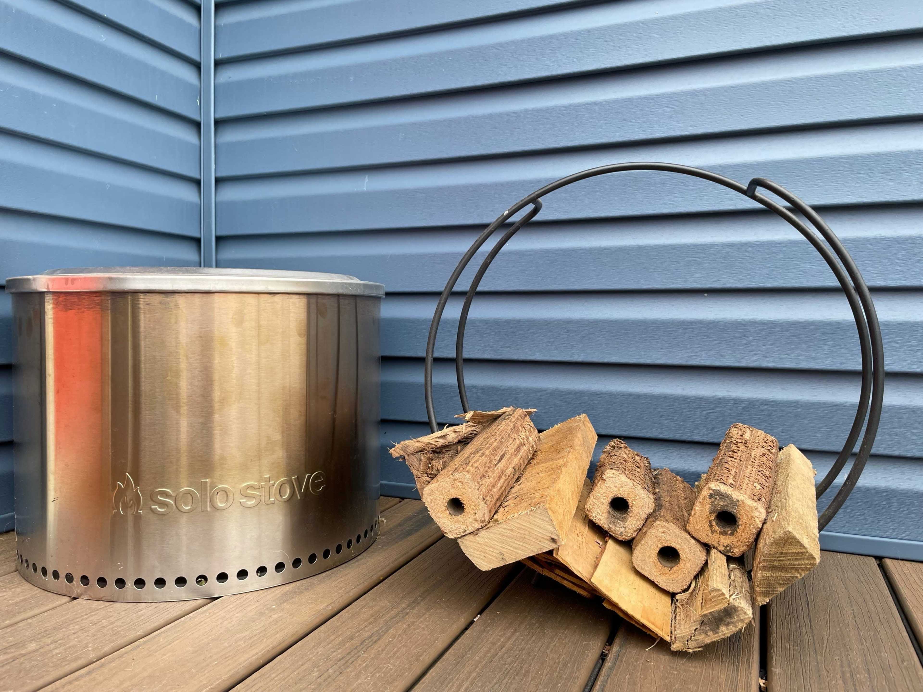 A stainless steel solo stove is positioned next to a metal wood holder filled with stacked firewood, all set against a blue wooden wall.