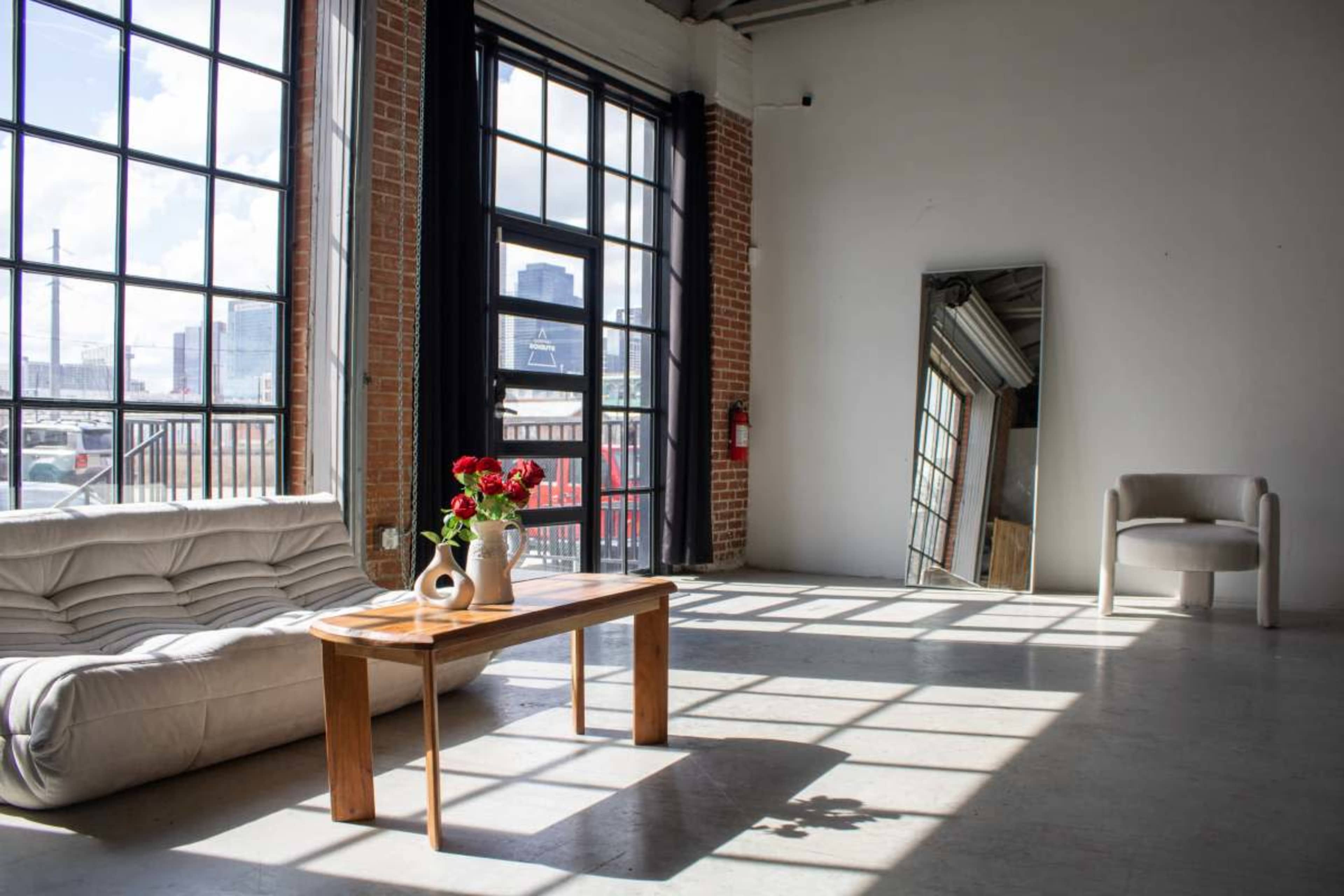 The image shows a bright, spacious room with large windows, a beige sofa, a wooden coffee table with a vase of flowers, a tall mirror, and a single chair.