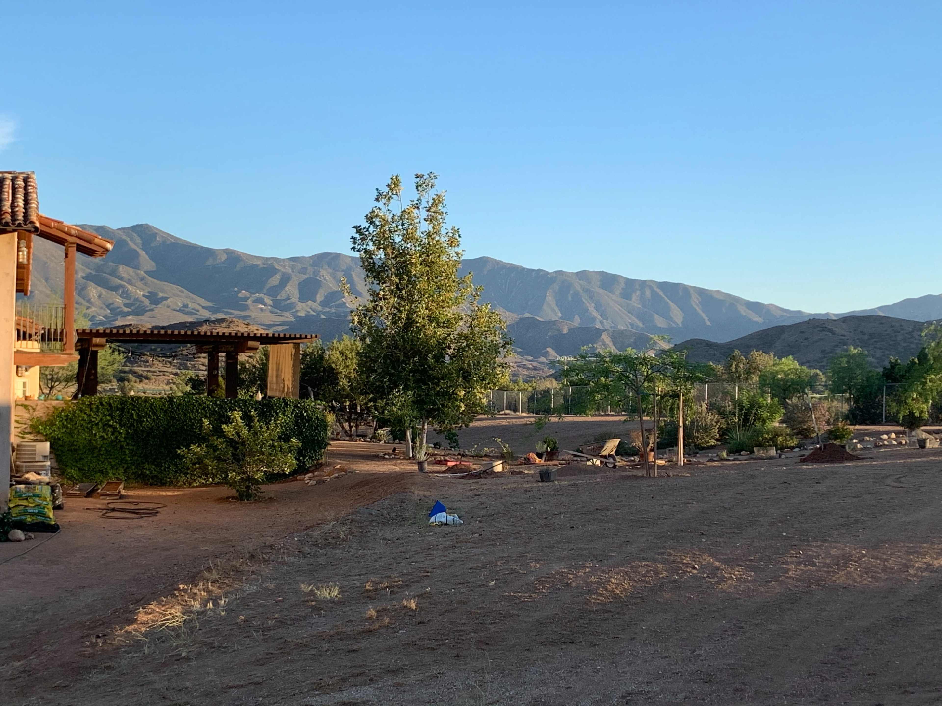 The scene shows a rural landscape with a house on the left, surrounded by sparse vegetation and mountains in the background under a clear sky.