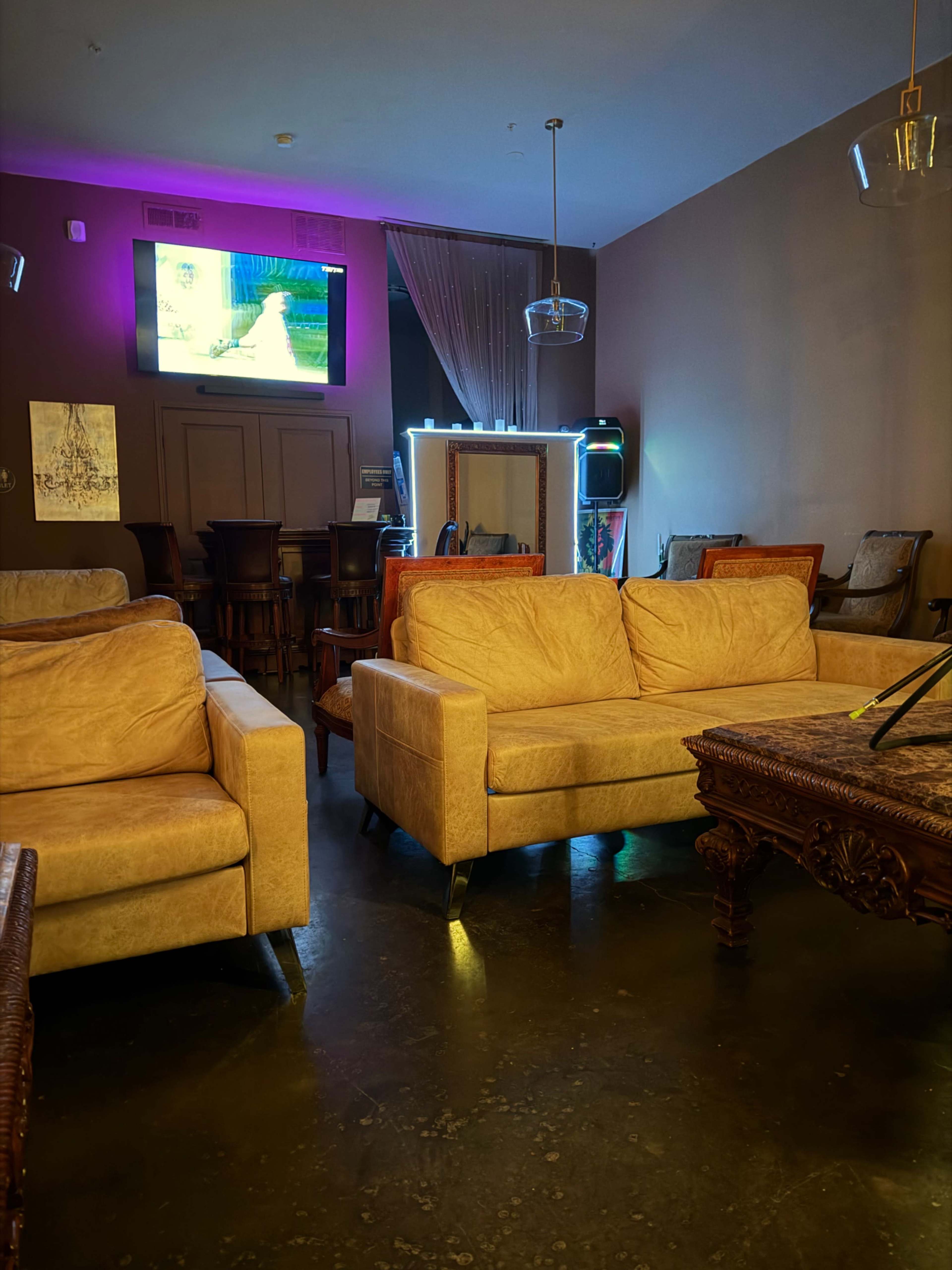 The image shows a cozy lounge area with yellow sofas, a wooden coffee table, and a dimly lit background featuring a television and a bar-like setting.