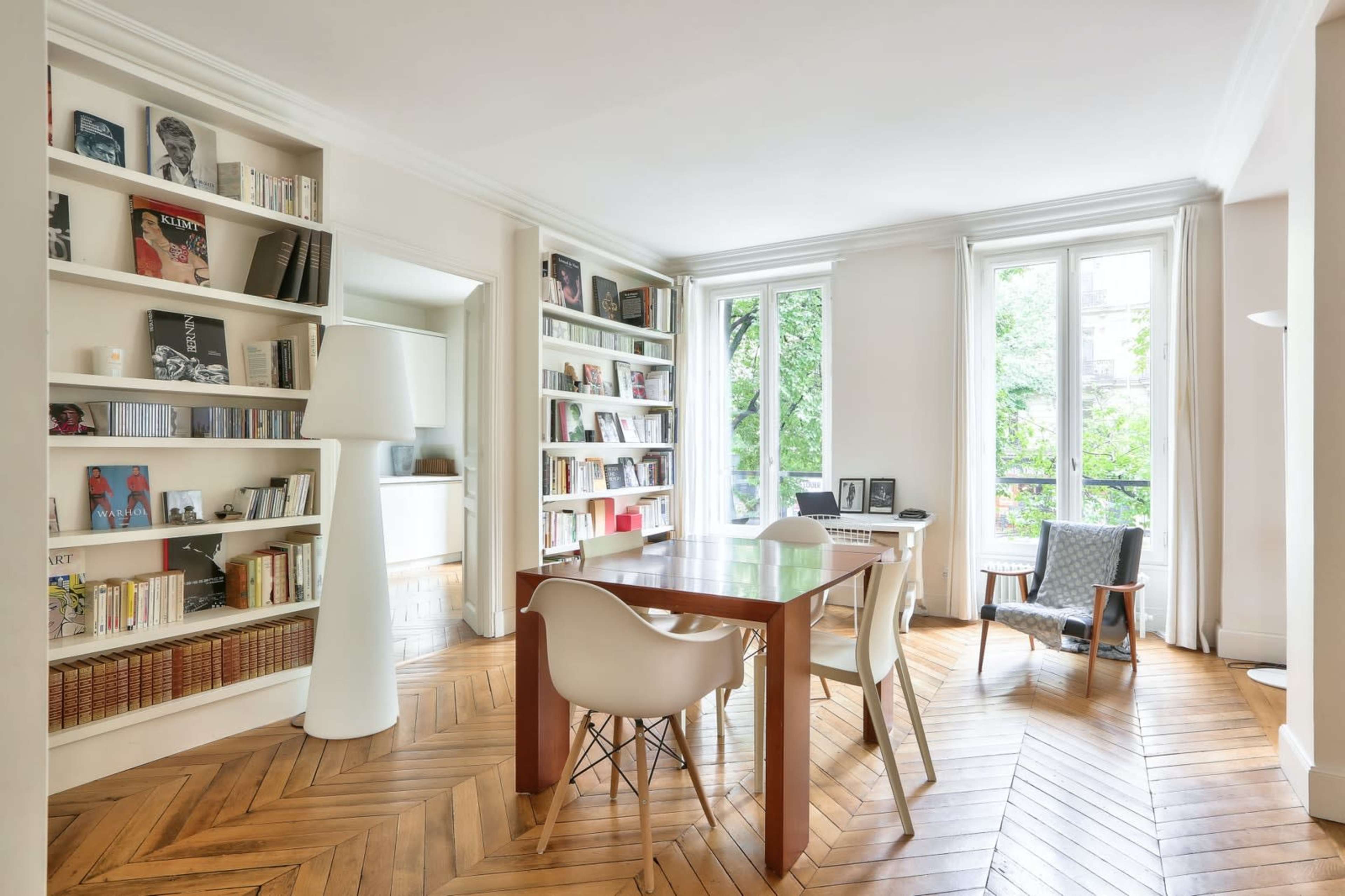 A bright room features a wooden dining table surrounded by modern chairs, with bookshelves lining the walls and large windows allowing natural light to enter.
