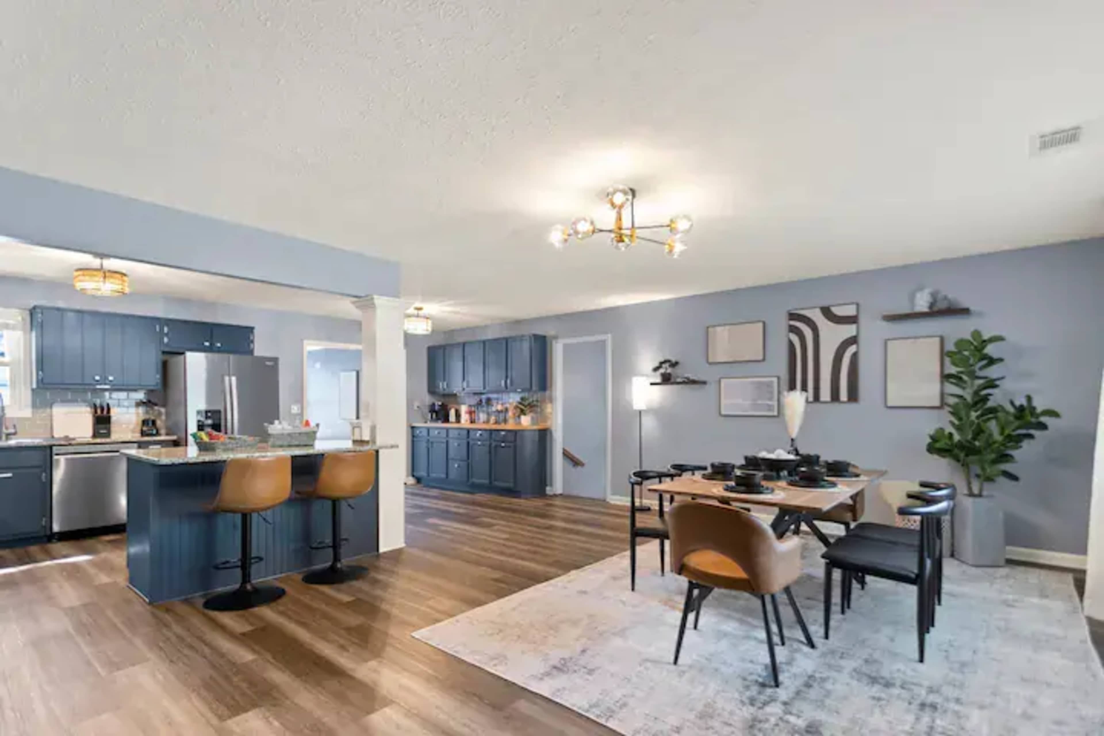 A modern kitchen and dining area feature blue cabinetry, a large island with bar stools, and a dining table set with dishware, all accented by wooden flooring and contemporary decor.