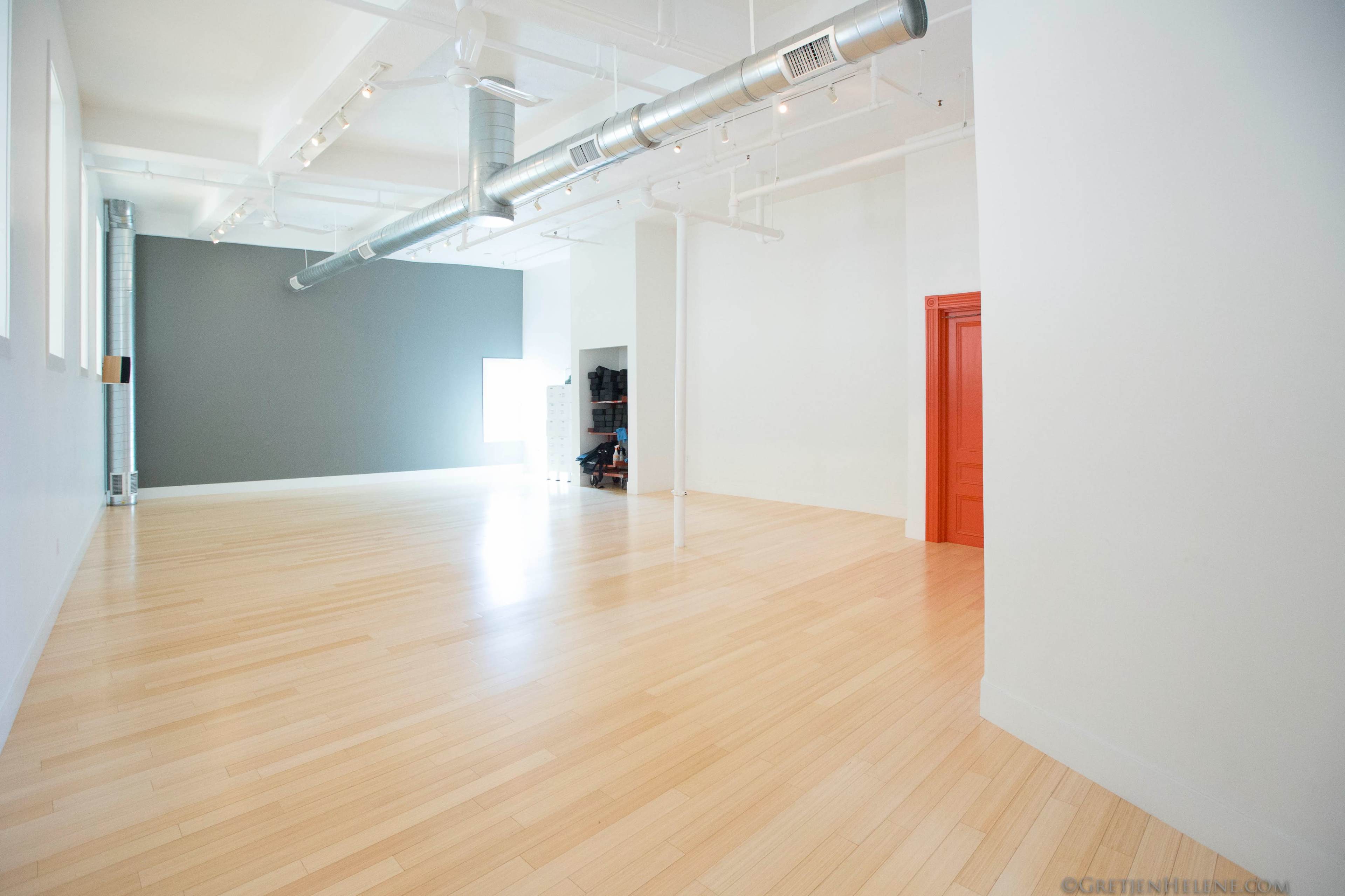 The image shows a spacious, empty room with light wood flooring, a gray accent wall, and a bright orange door.