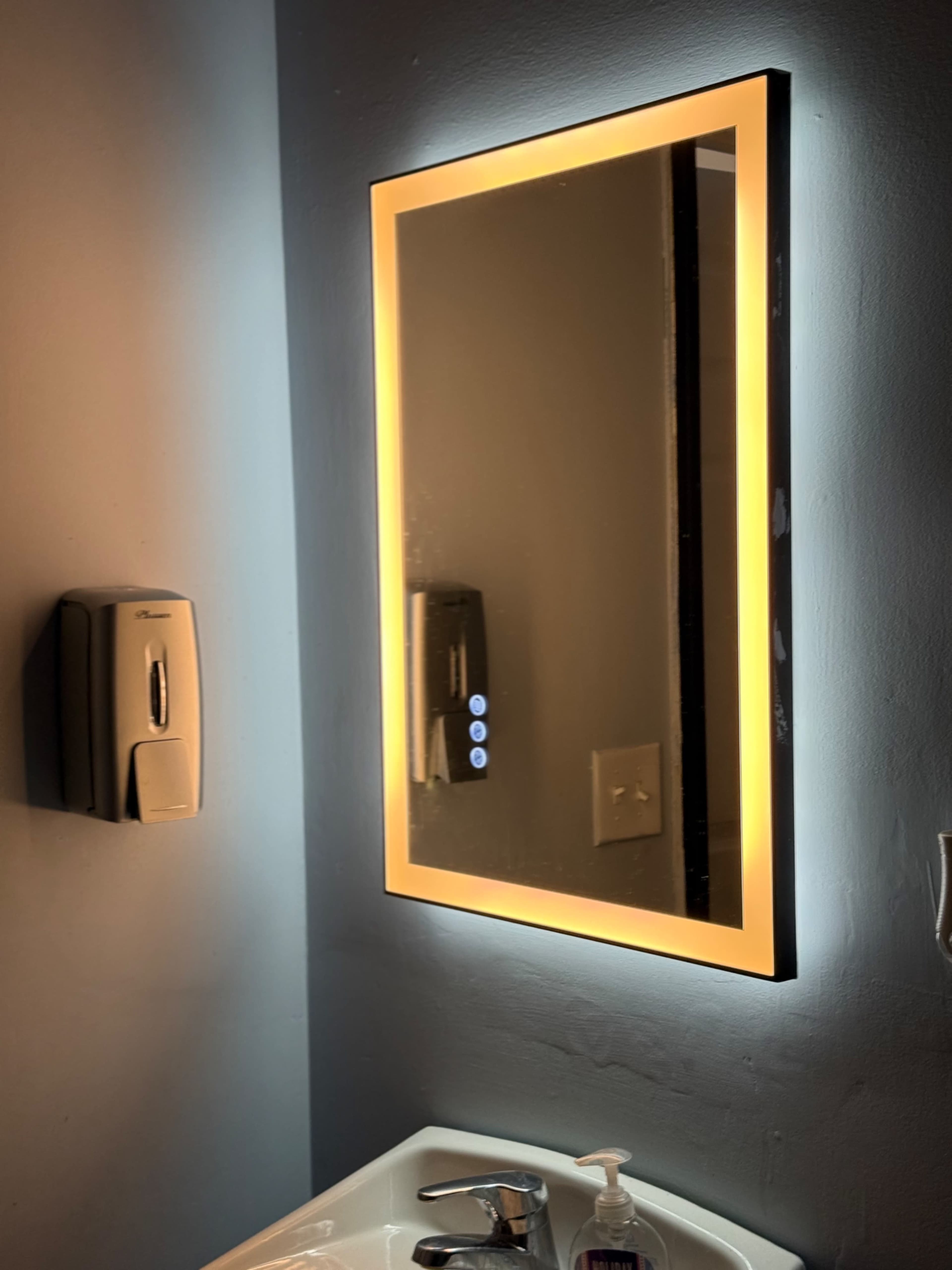 The image shows a bathroom with a backlit mirror above a sink, accompanied by a soap dispenser mounted on the wall.