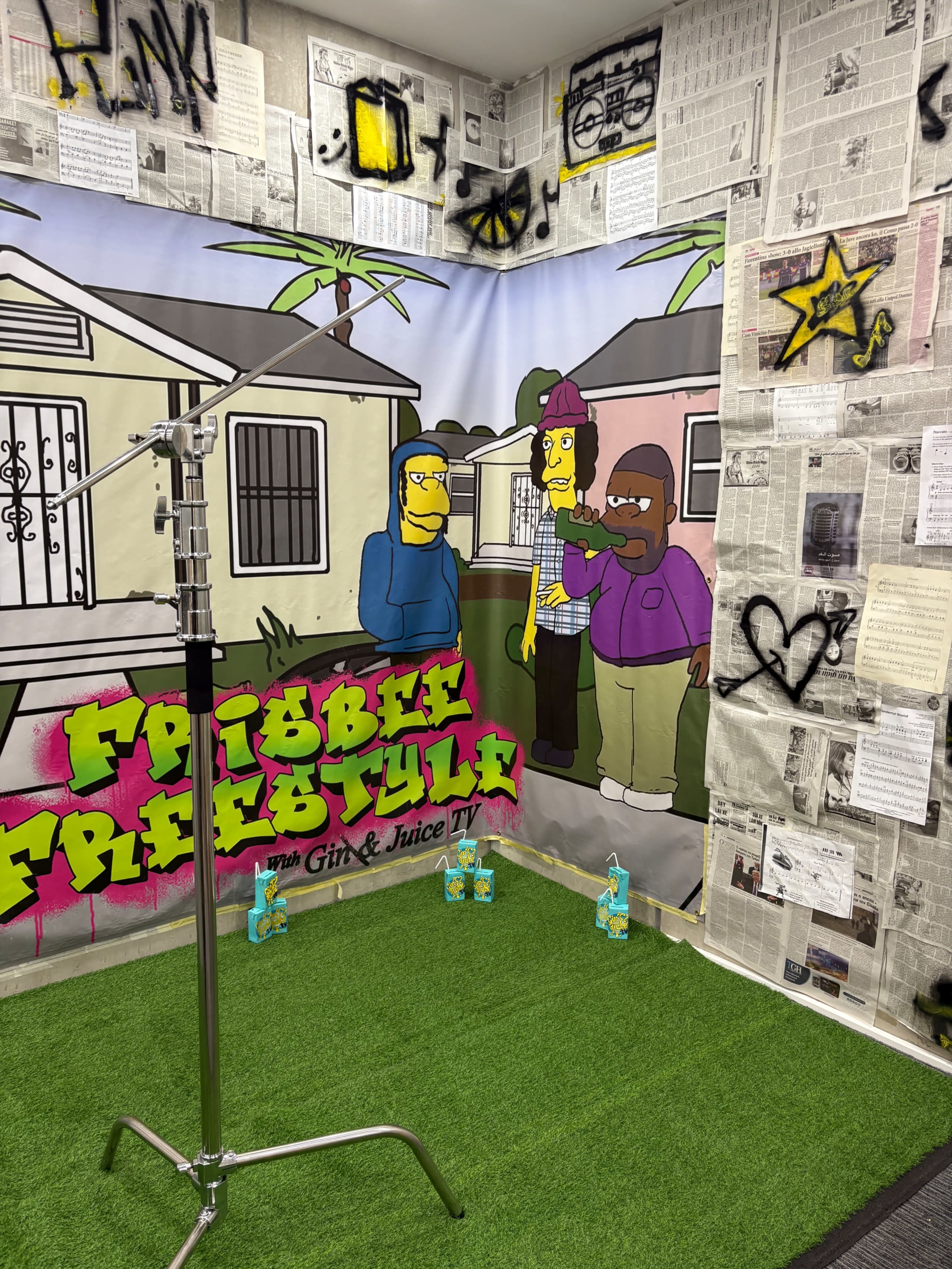 The image features a colorful backdrop depicting cartoon characters near a house, with newspaper clippings and graffiti on the walls in a small studio setting.