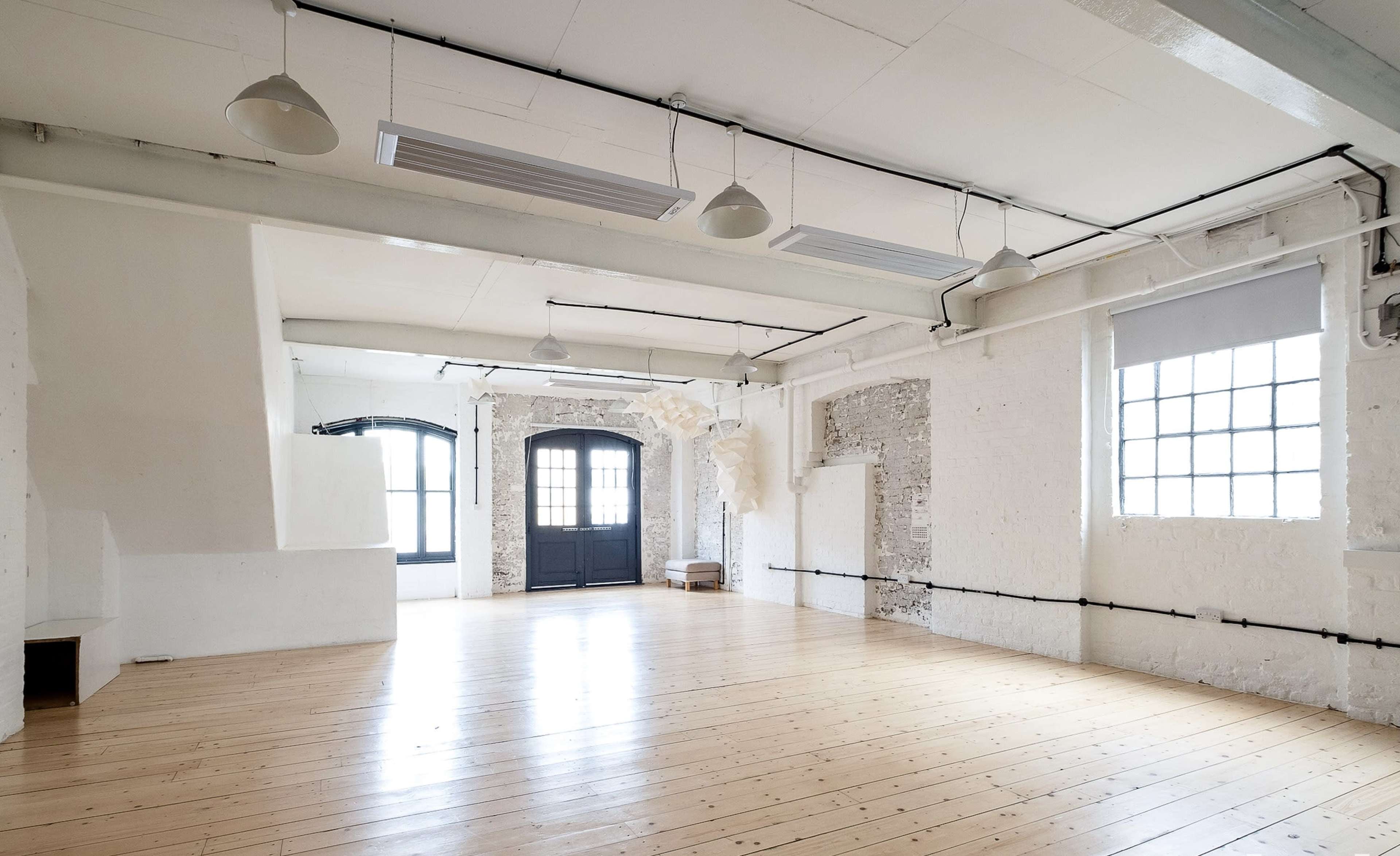 A spacious, empty room with wooden flooring, white walls, large windows, and industrial-style light fixtures.