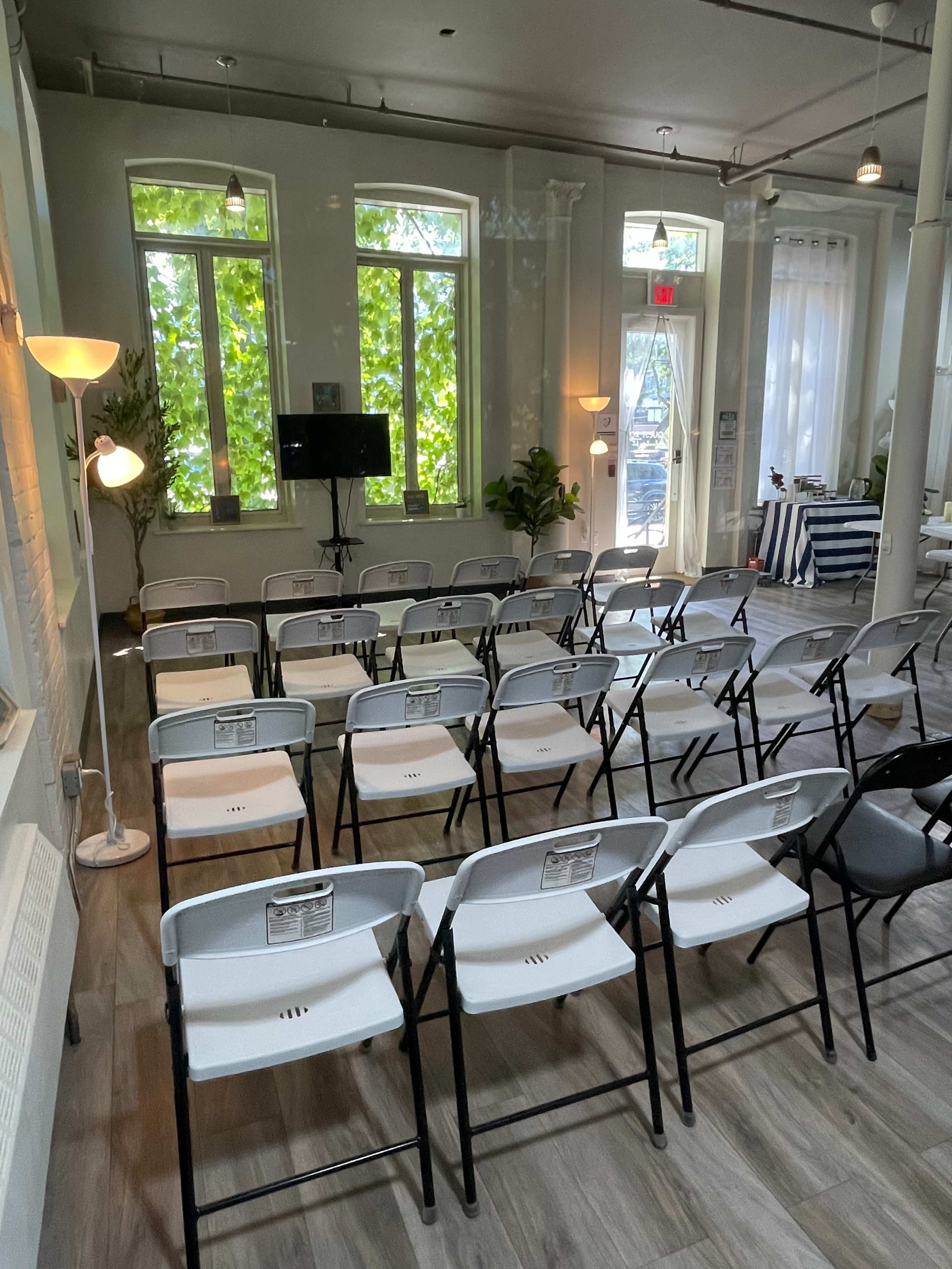 A room arranged with rows of white folding chairs facing a TV screen, with large windows allowing sunlight to enter.