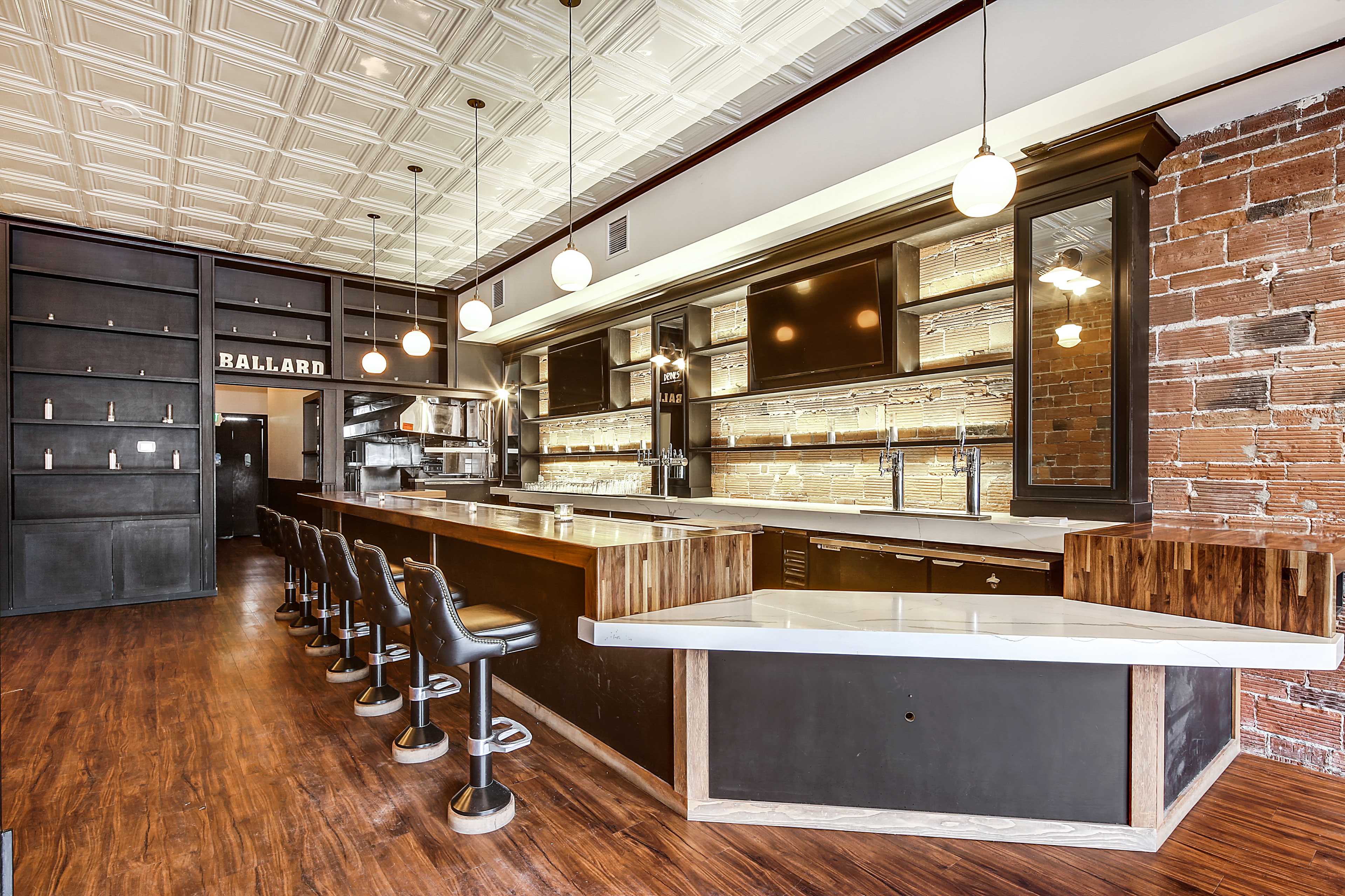 A modern bar with a long counter, wooden accents, and pendant lighting, positioned against a backdrop of exposed brick and a coffered ceiling.