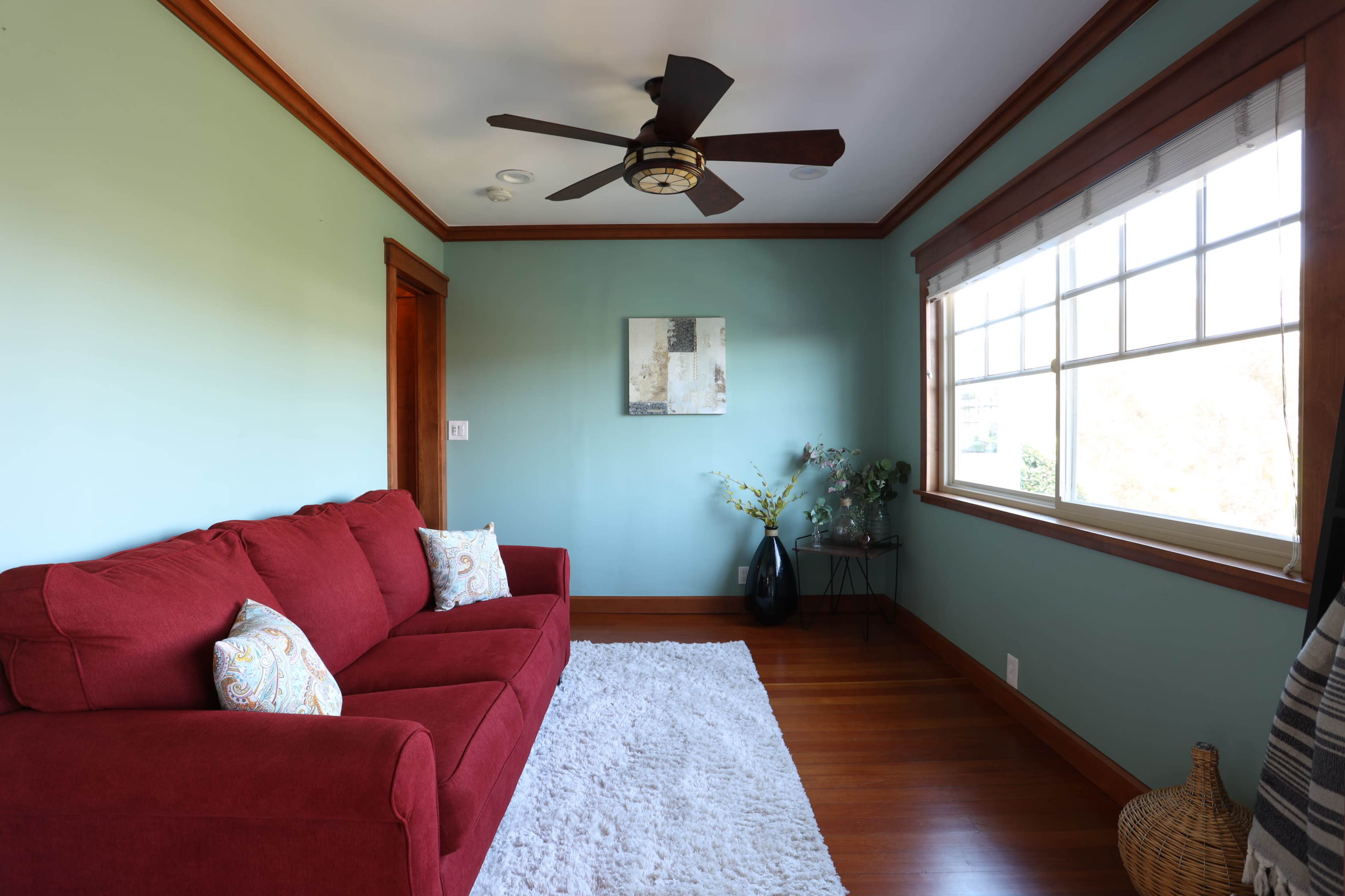 The image shows a bright room with a red sofa, a light rug on hardwood flooring, and a window with wooden trim.