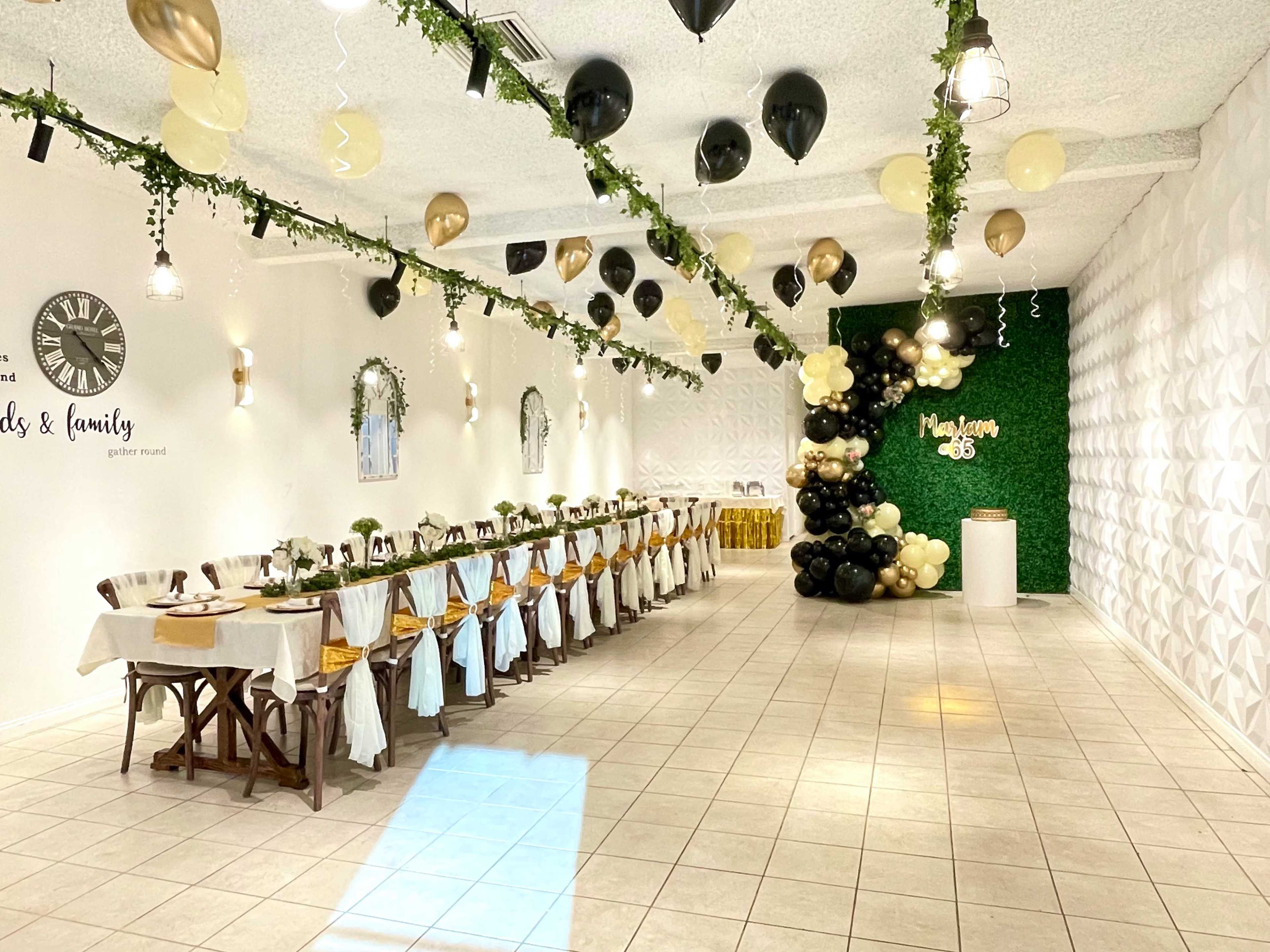 A banquet hall decorated with green foliage, black and gold balloons, and a long dining table set for an event.