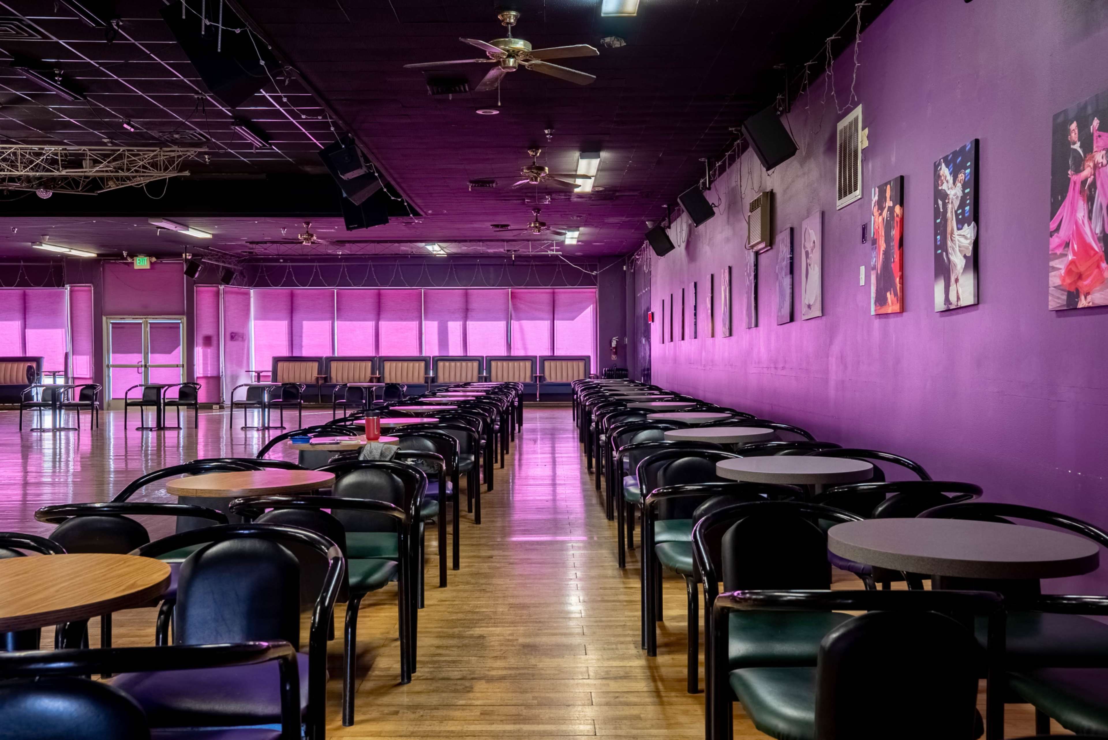 The image shows an expansive dance hall with rows of black chairs and tables, illuminated by pink lighting and adorned with framed photographs on the walls.