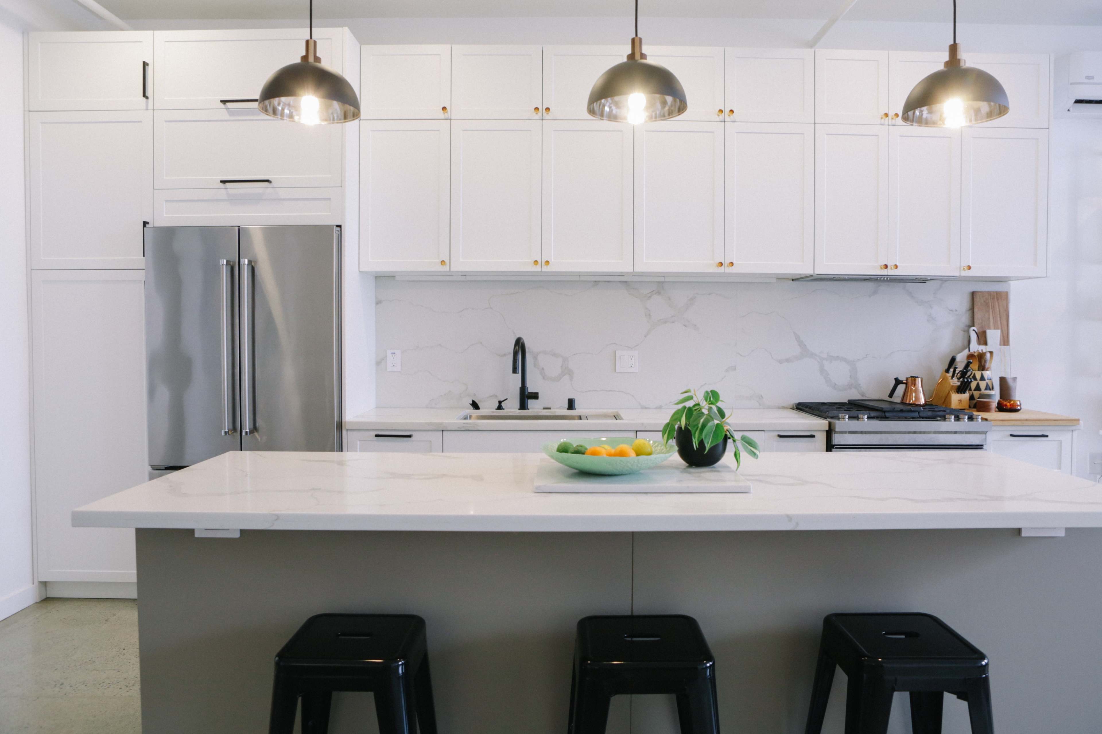 A modern kitchen features white cabinets, a marble countertop, a stainless steel fridge, and pendant lights above a central island.