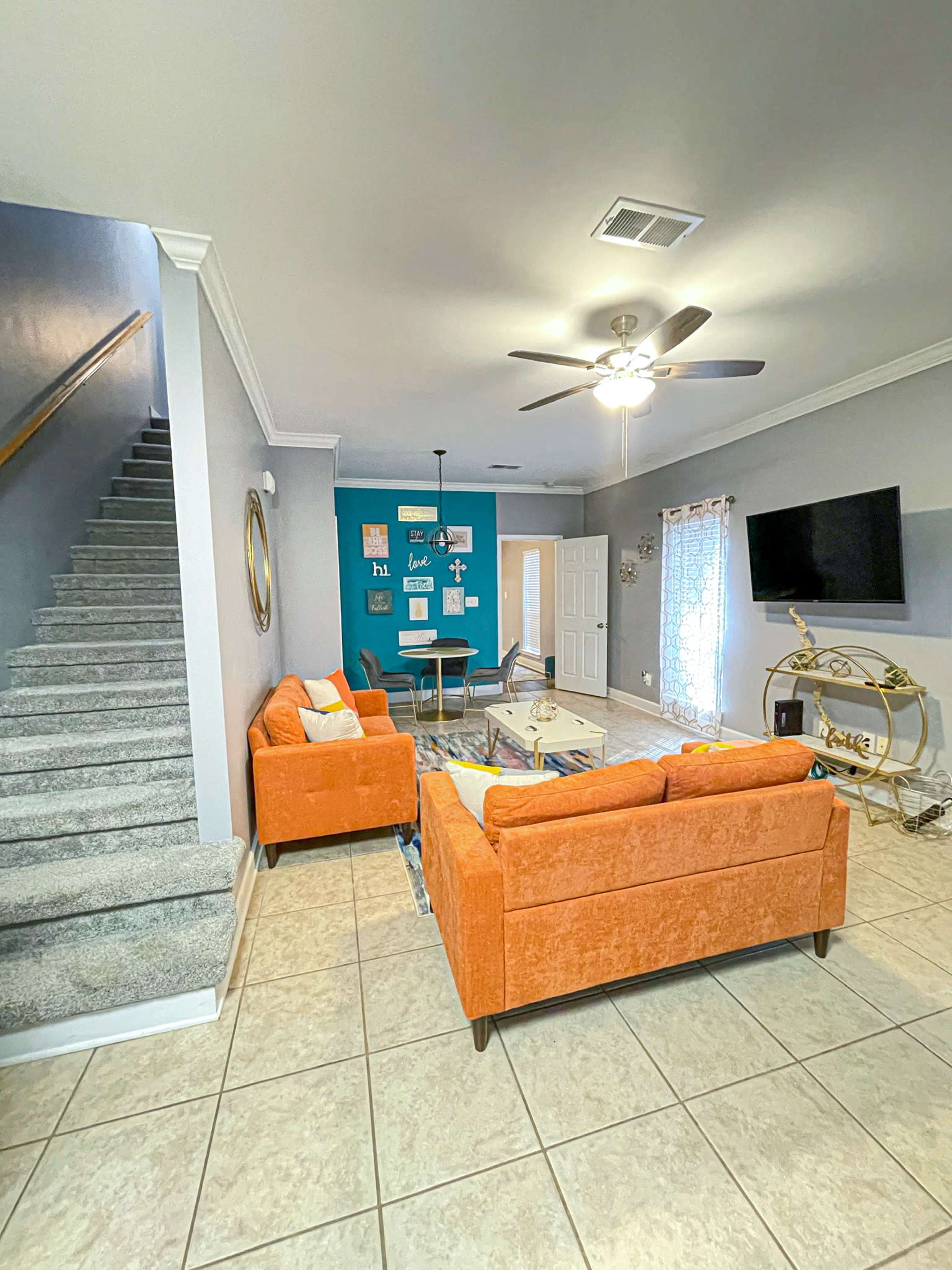 The living room features orange couches, a coffee table, and a television against a blue accent wall, with stairs leading up on one side.