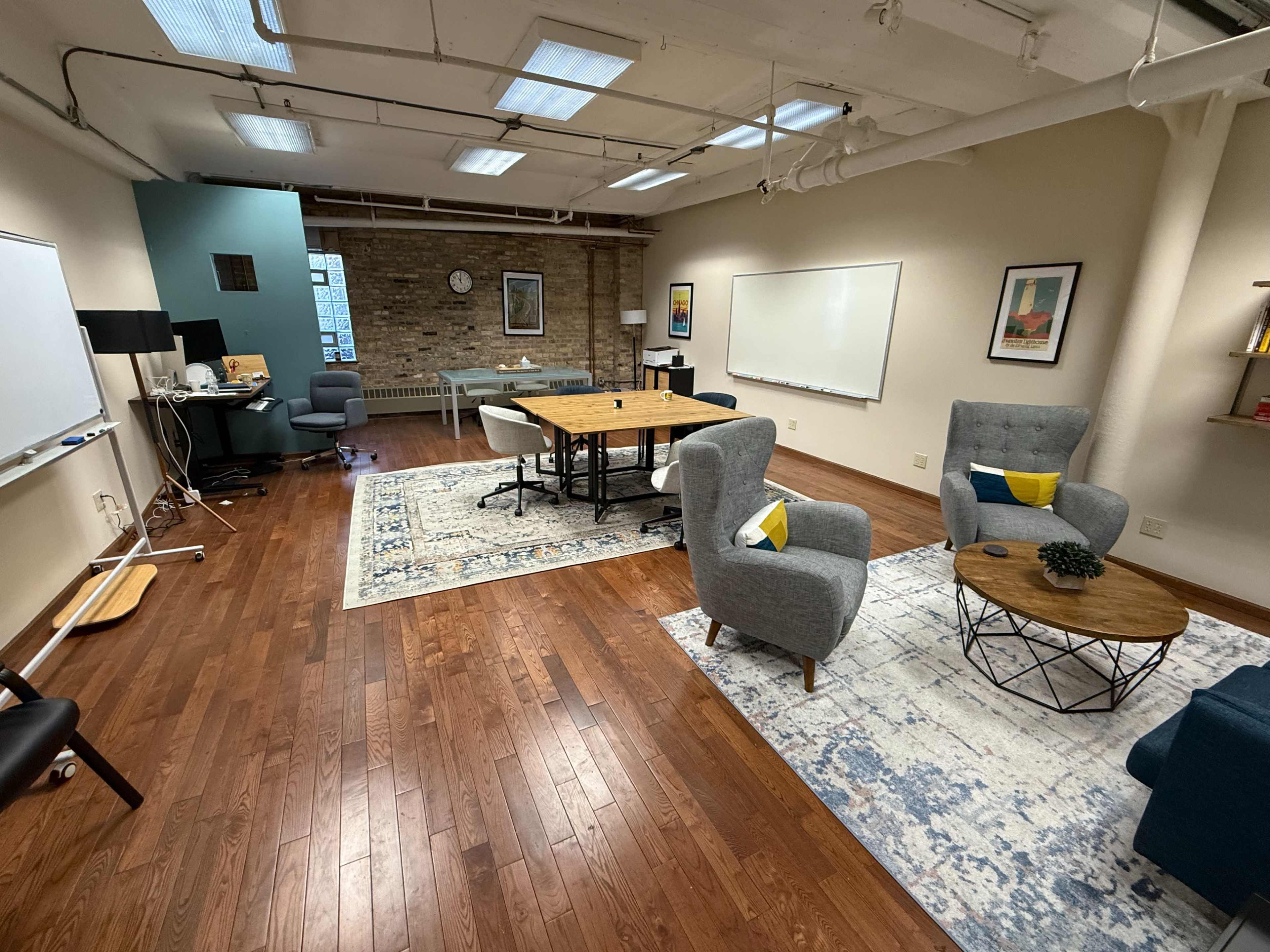 The image shows a spacious office with wooden floors, a large table surrounded by chairs, and cozy seating areas, featuring a whiteboard and a large window in the background.